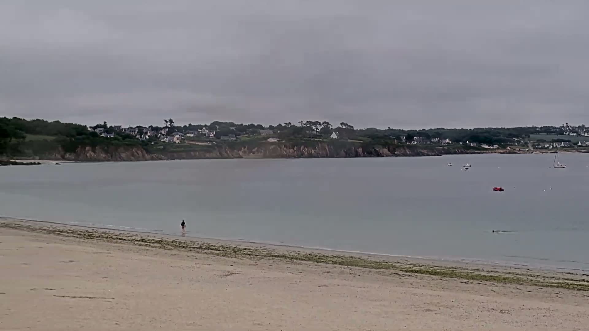 A person walks along a sandy beach toward calm, light-grey water under an overcast sky, with a few boats visible in the distance and a small village nestled on the far shore.
