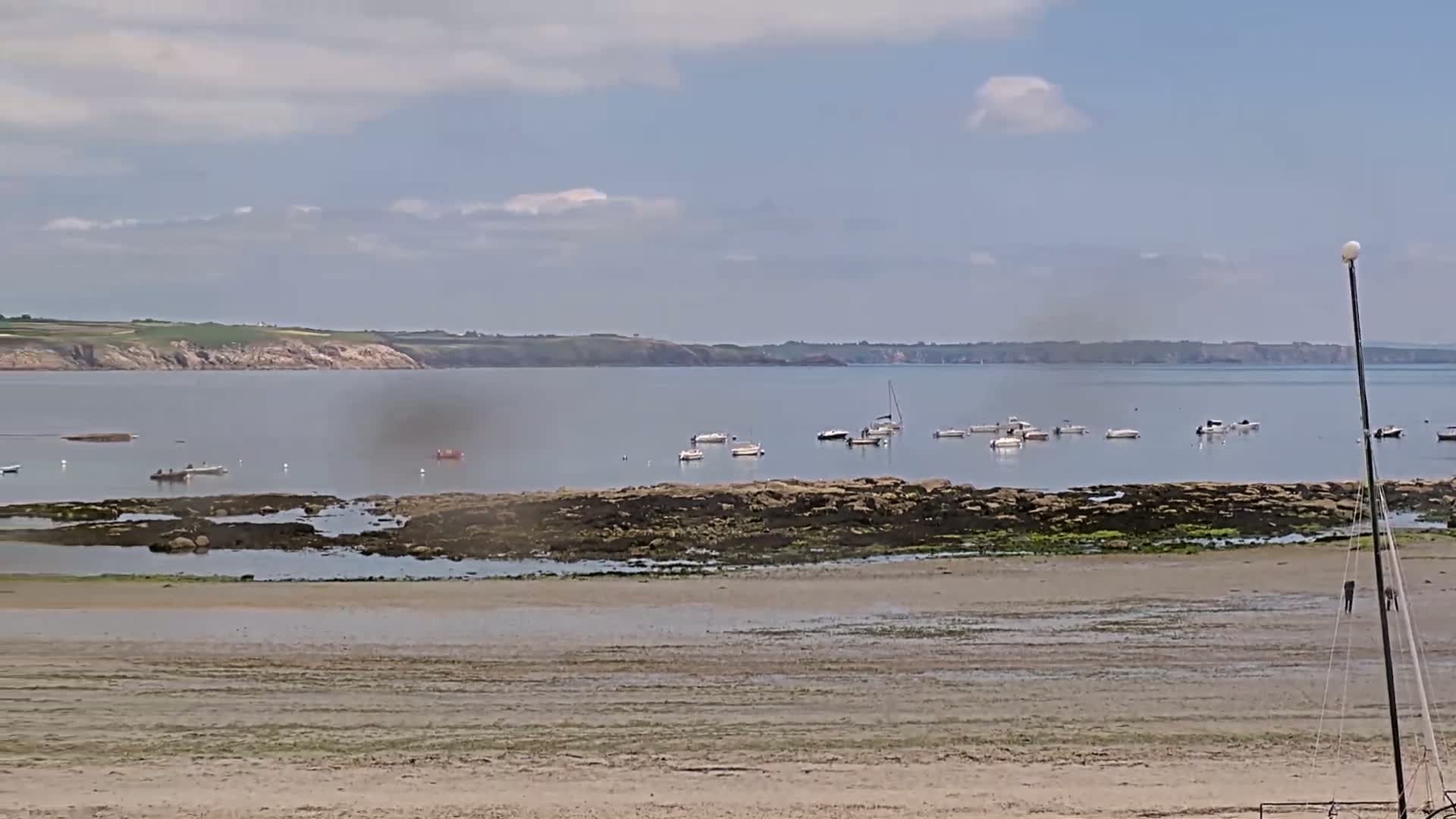 A sandy beach stretches to a calm bay dotted with several small boats, under a partly cloudy sky.