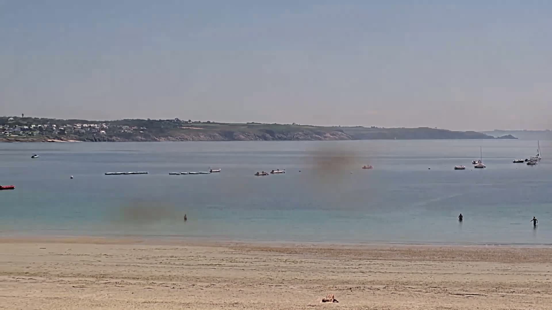 A sandy beach overlooks a calm, clear bay with several small boats and people swimming, under a hazy sunny sky.