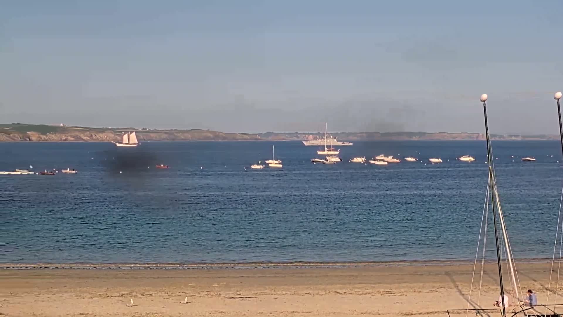 A sandy beach overlooks a calm sea where several small boats and two larger vessels are anchored, with a distant shoreline visible under a clear sky.