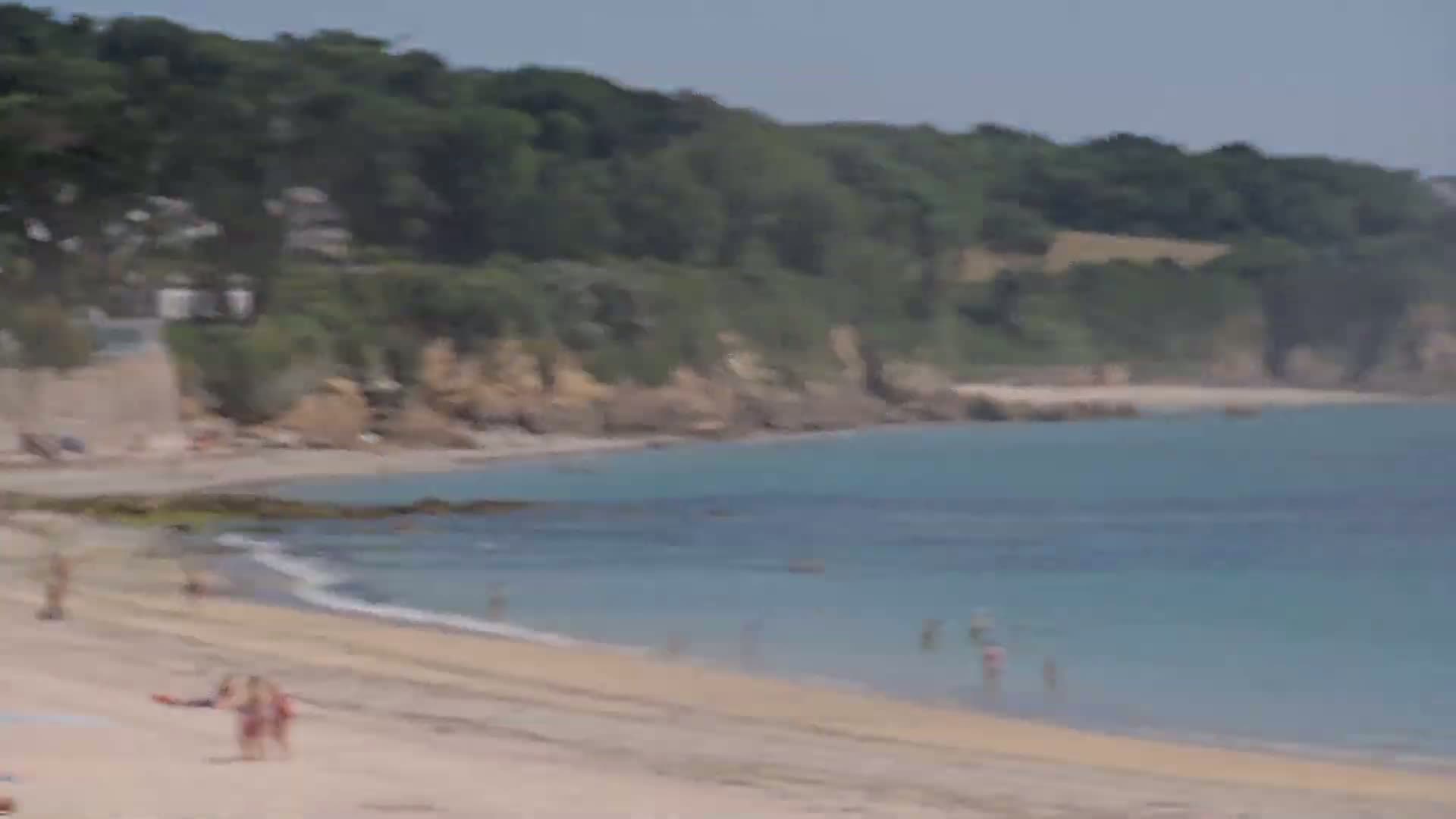 A sandy beach with a few people on it is bordered by calm blue water and a green, hilly coastline on a sunny day.
