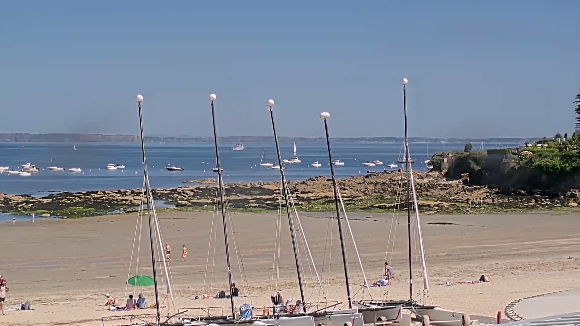 Several sailboats are beached on a sandy shore, with people relaxing nearby under a clear, sunny sky and a calm body of water dotted with other boats in the distance.