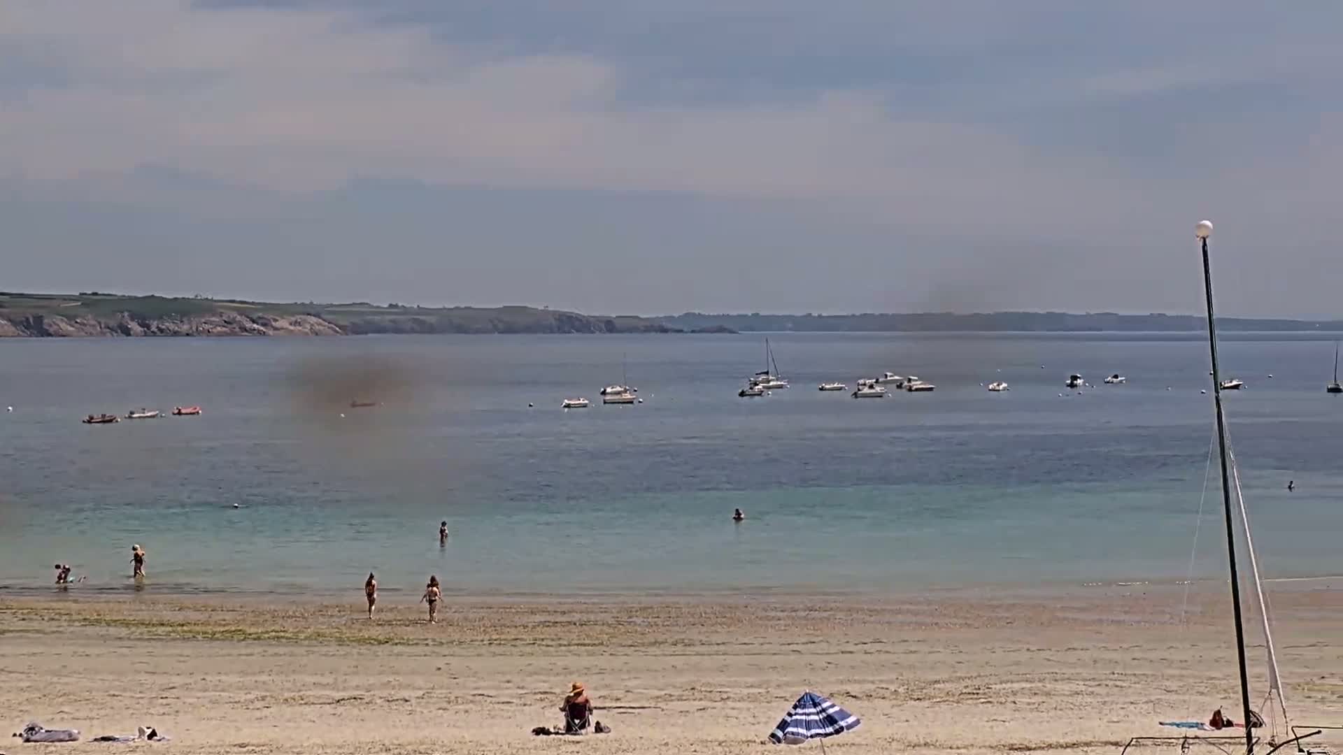 A sandy beach with several people swimming and sunbathing, boats on calm, light-blue water, and a hazy sky.
