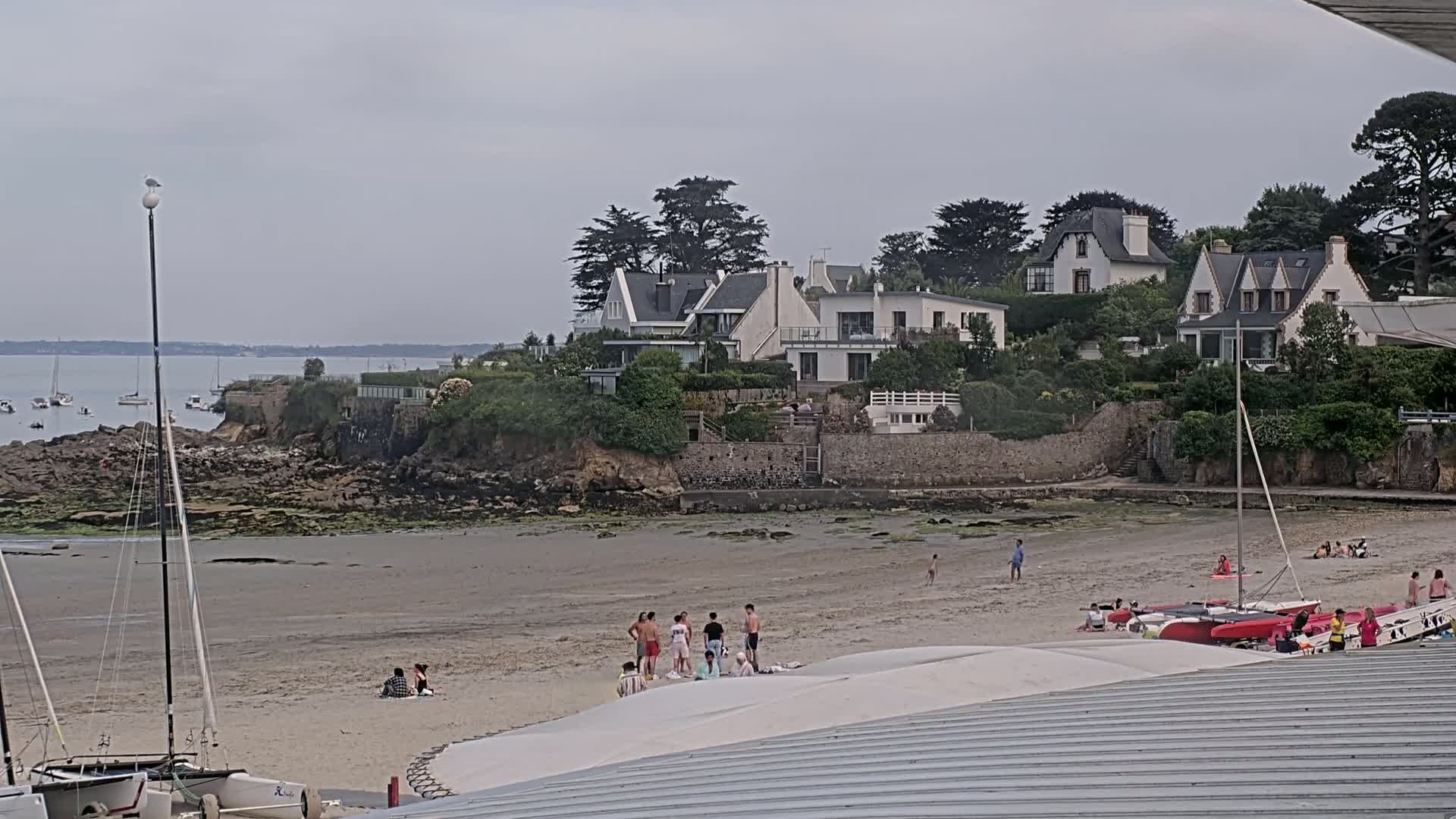 A sandy beach with several people, sailboats, and houses on a hill overlooking the water under an overcast sky.