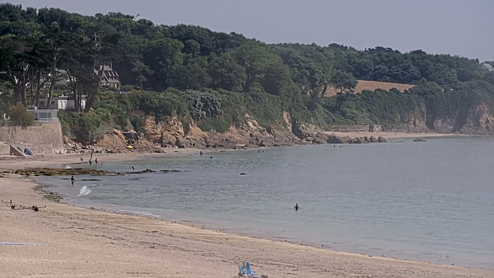A sandy beach with calm, light-blue water stretches out toward a rocky coastline covered in lush green trees and houses under a hazy, sunny sky.