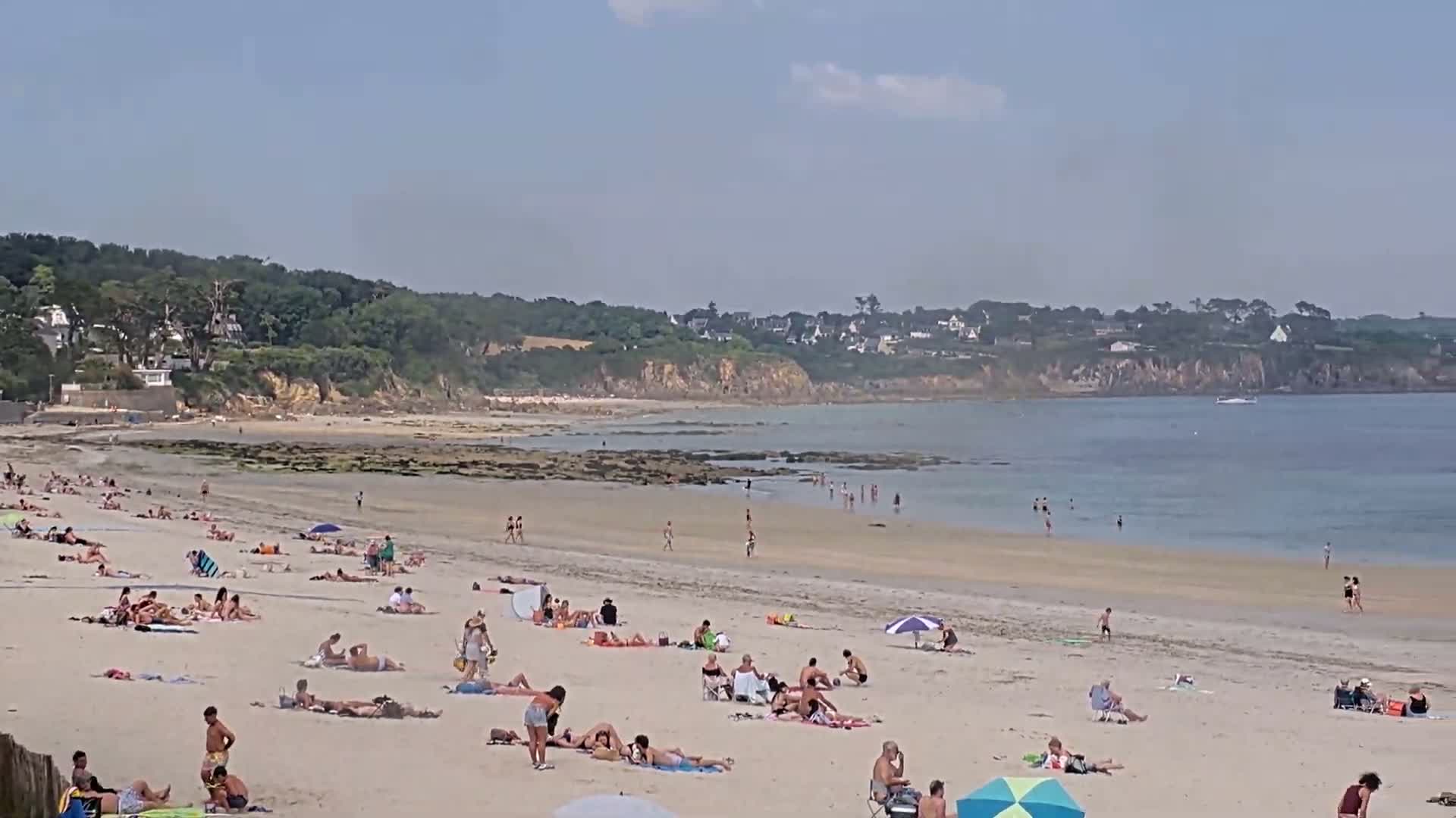 A sandy beach with many people sunbathing and swimming in calm, shallow water on a sunny day, backed by green hills and houses.