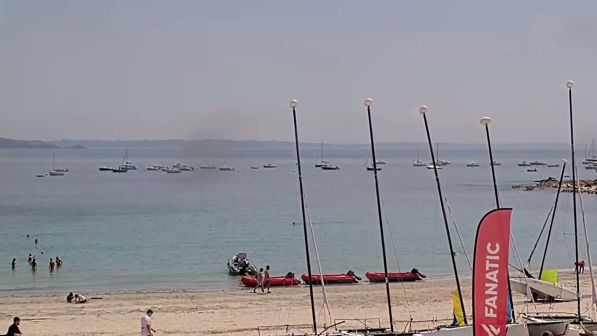 A sandy beach with several people swimming and walking, numerous boats on the calm, light-blue water, and sailboats moored on the shore under a hazy, sunny sky.