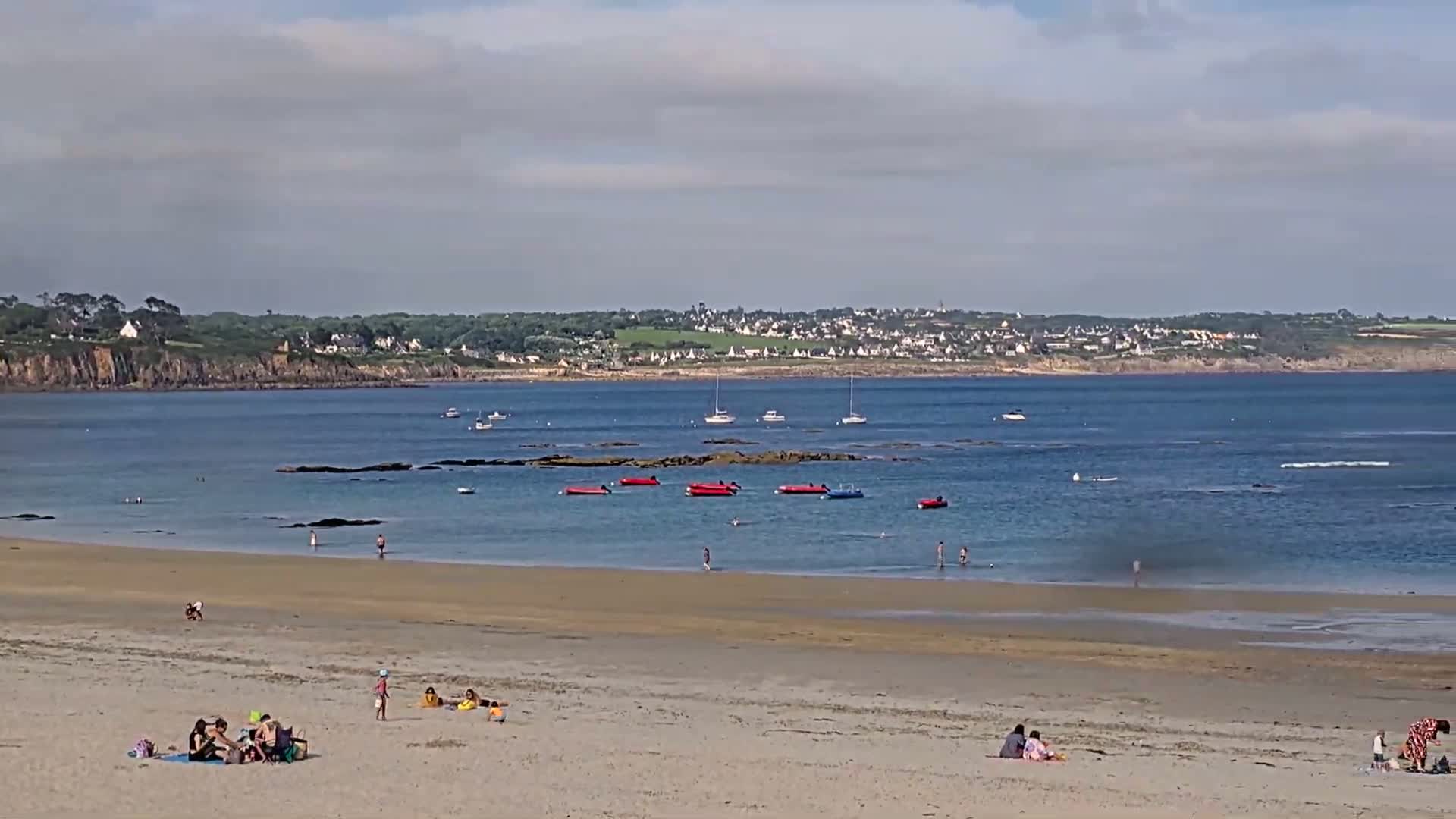 A sandy beach with people sunbathing and swimming in calm water, several small boats anchored offshore, and a town visible in the distance under a partly cloudy sky.