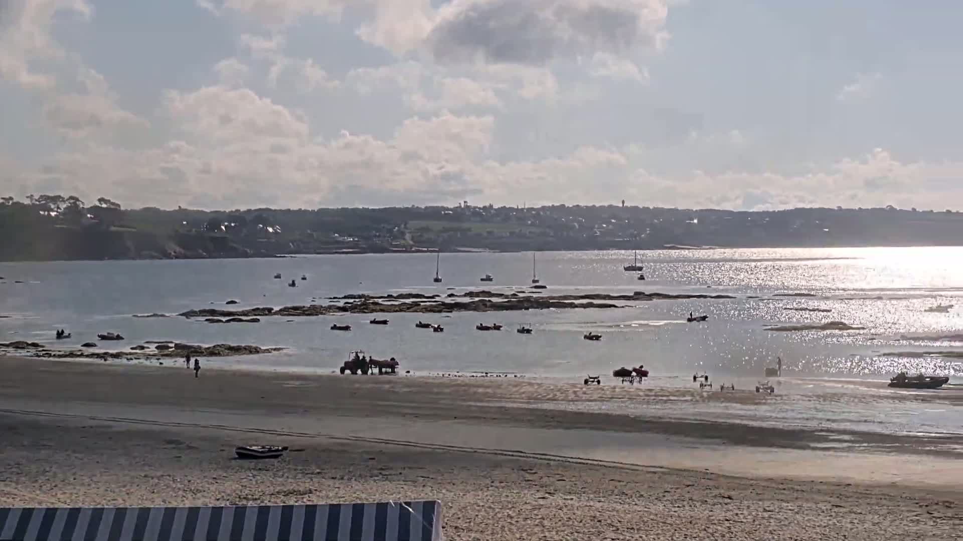 A sandy beach with several small boats and people near the water's edge, under a partly cloudy sky, with a calm, sparkling sea and distant shoreline visible.
