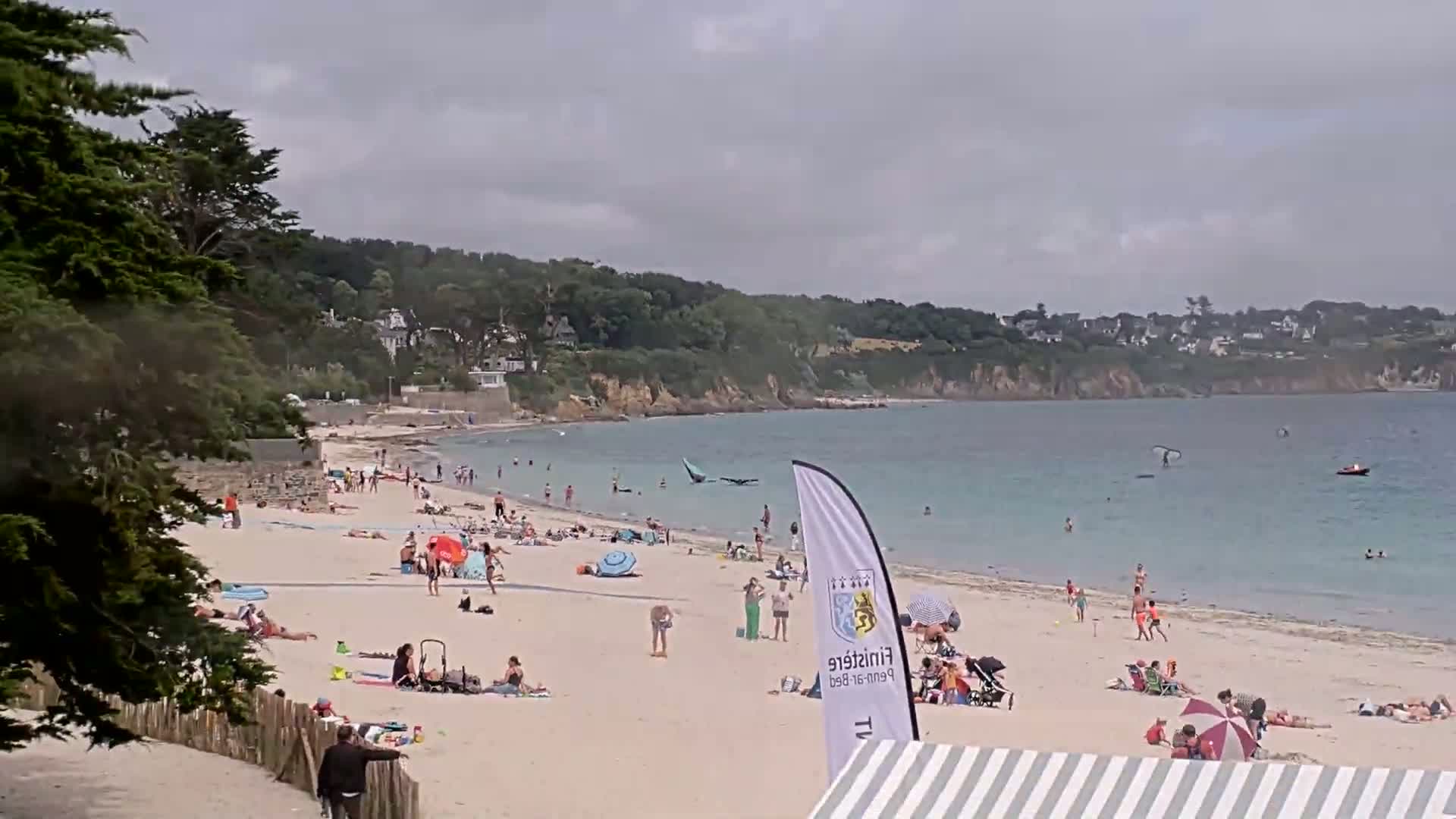 A sandy beach with many people relaxing and swimming in calm, light-blue water under an overcast sky, backed by green hills and houses.