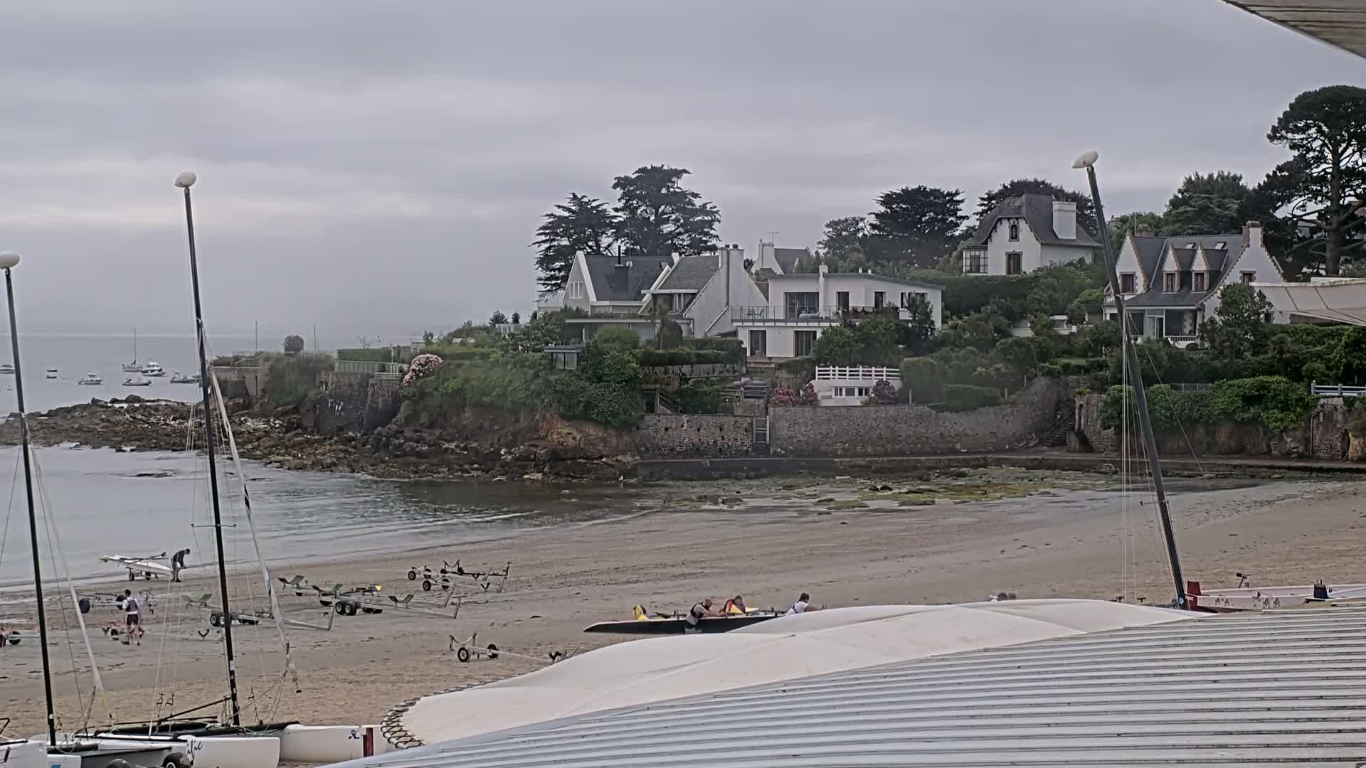 A sandy beach with several sailboats and their trailers is in the foreground, with a cluster of white houses on a small rocky peninsula under an overcast sky.