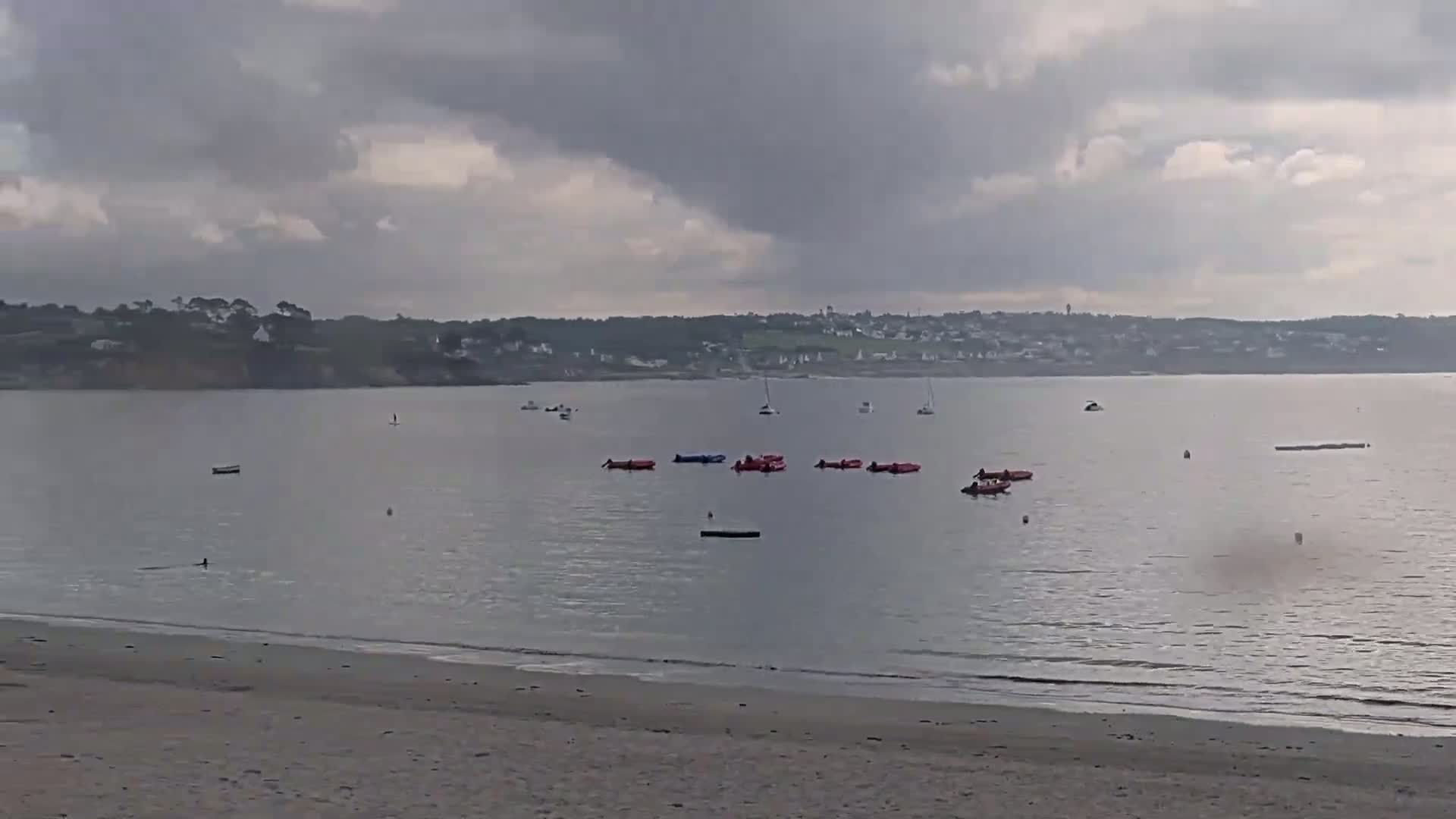 A sandy beach overlooks a calm bay under a cloudy sky, where several small boats and inflatable rafts are scattered on the water.
