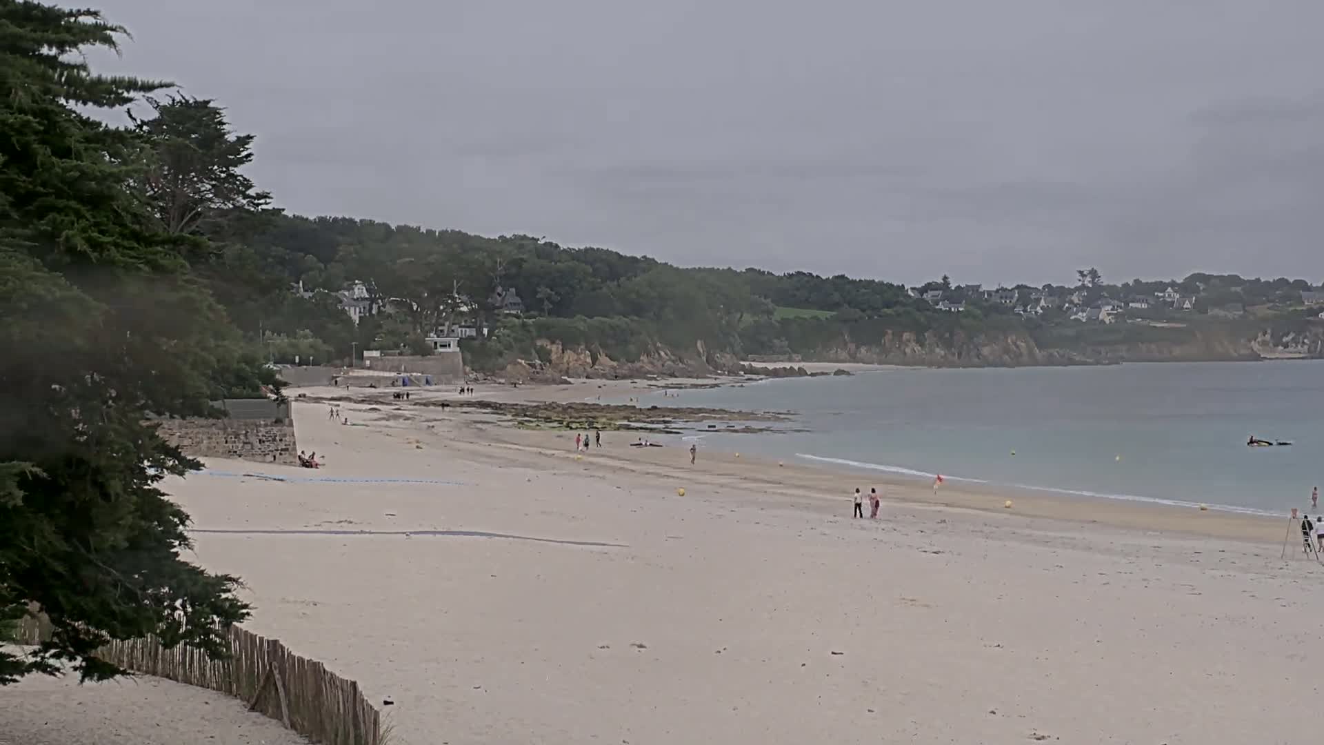 A mostly sunny day shows a wide sandy beach with a few people scattered across it,  backed by a rocky coastline and green trees, leading to a calm, light blue ocean.