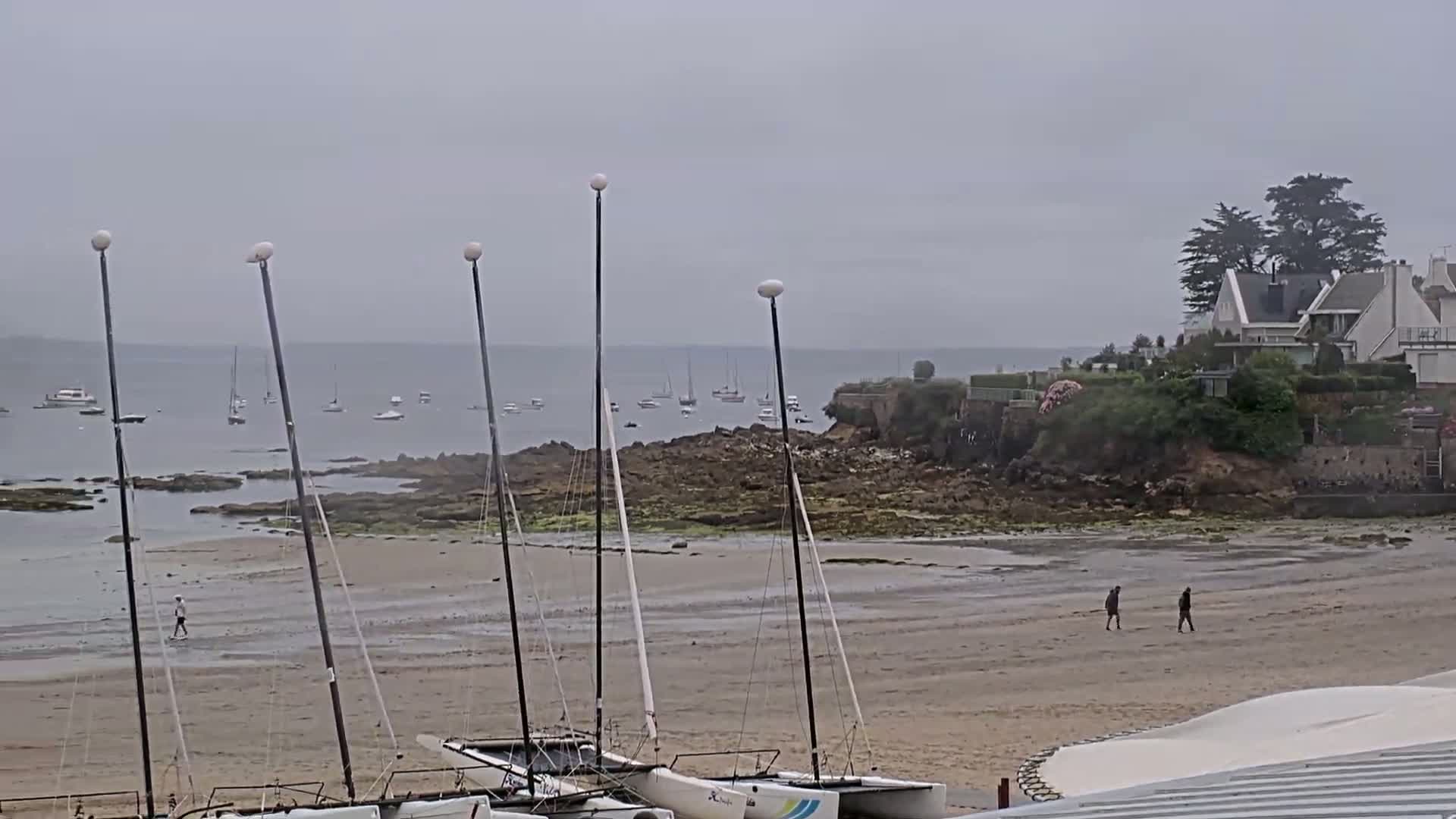 Several sailboats are beached on a sandy shore near rocky outcrops, with a calm sea and overcast skies in the background, and a few people walking on the beach.