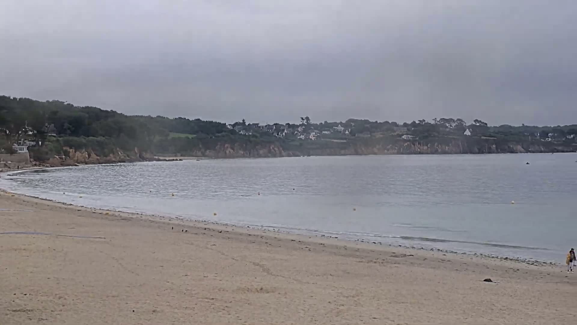 A sandy beach stretches towards a calm bay under an overcast sky, with a small village nestled on the hillside across the water.