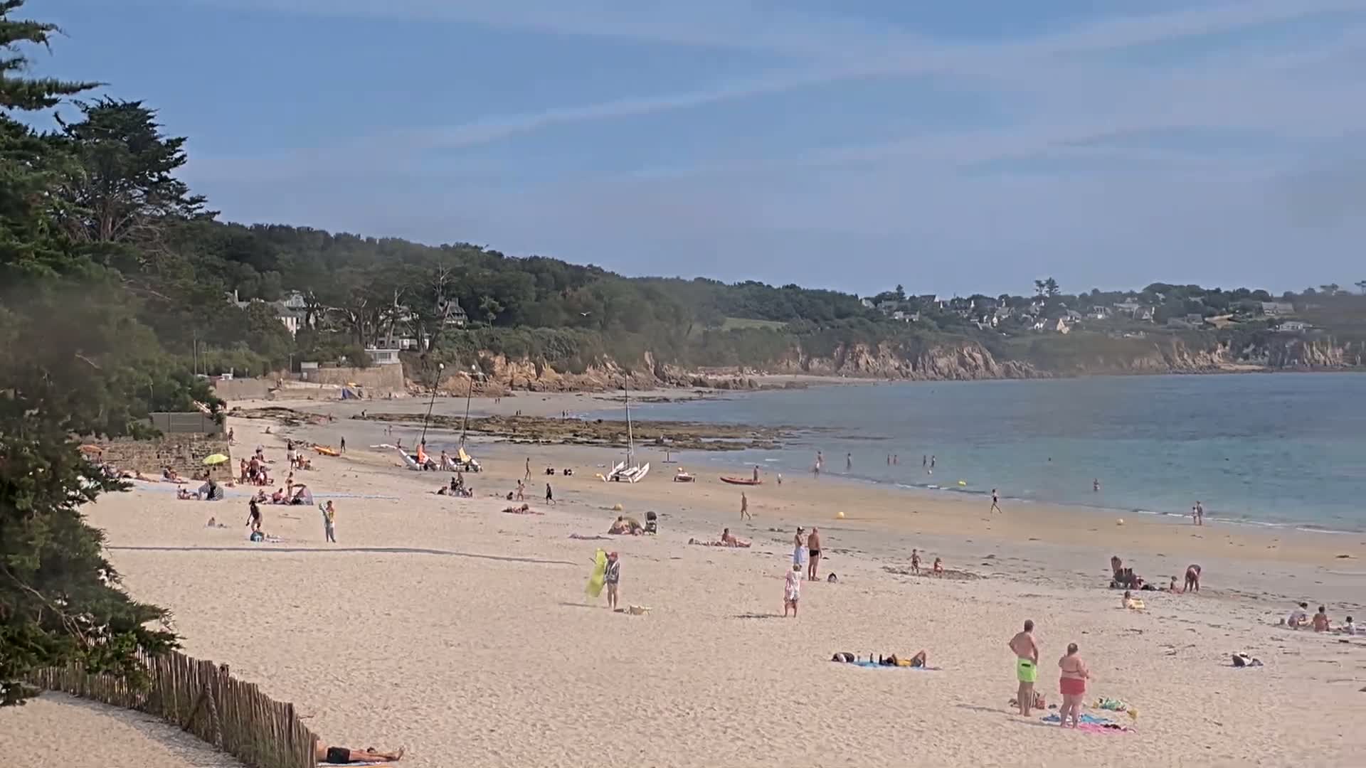 A sandy beach stretches towards a calm bay under an overcast sky, with a small village nestled on the hillside across the water.