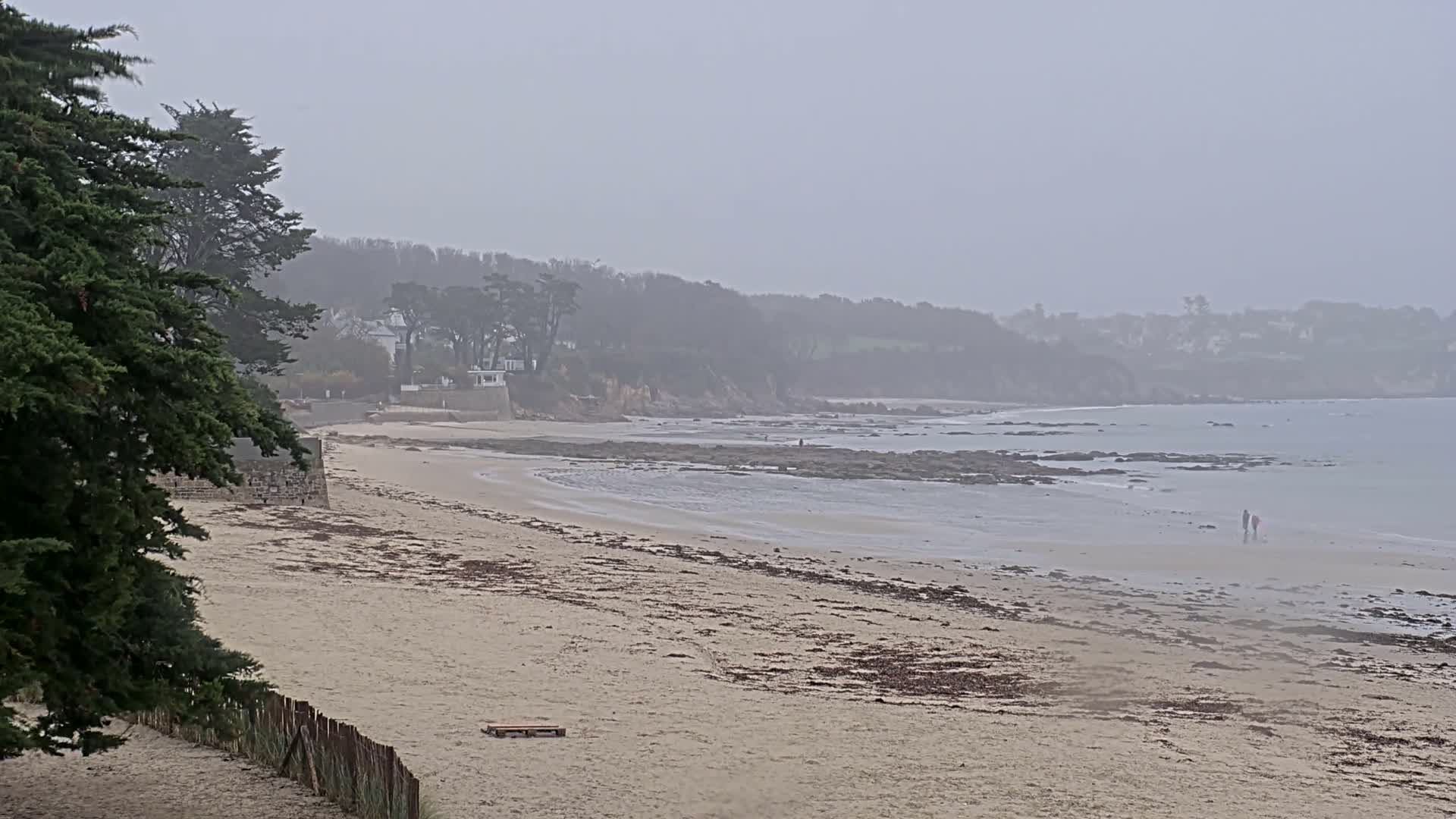 A wide sandy beach, scattered with seaweed and a few small structures, stretches along a rocky coastline with calm waters, backed by mist-shrouded, tree-covered hills and distant houses under an overcast sky, with two figures walking by the water's edge.