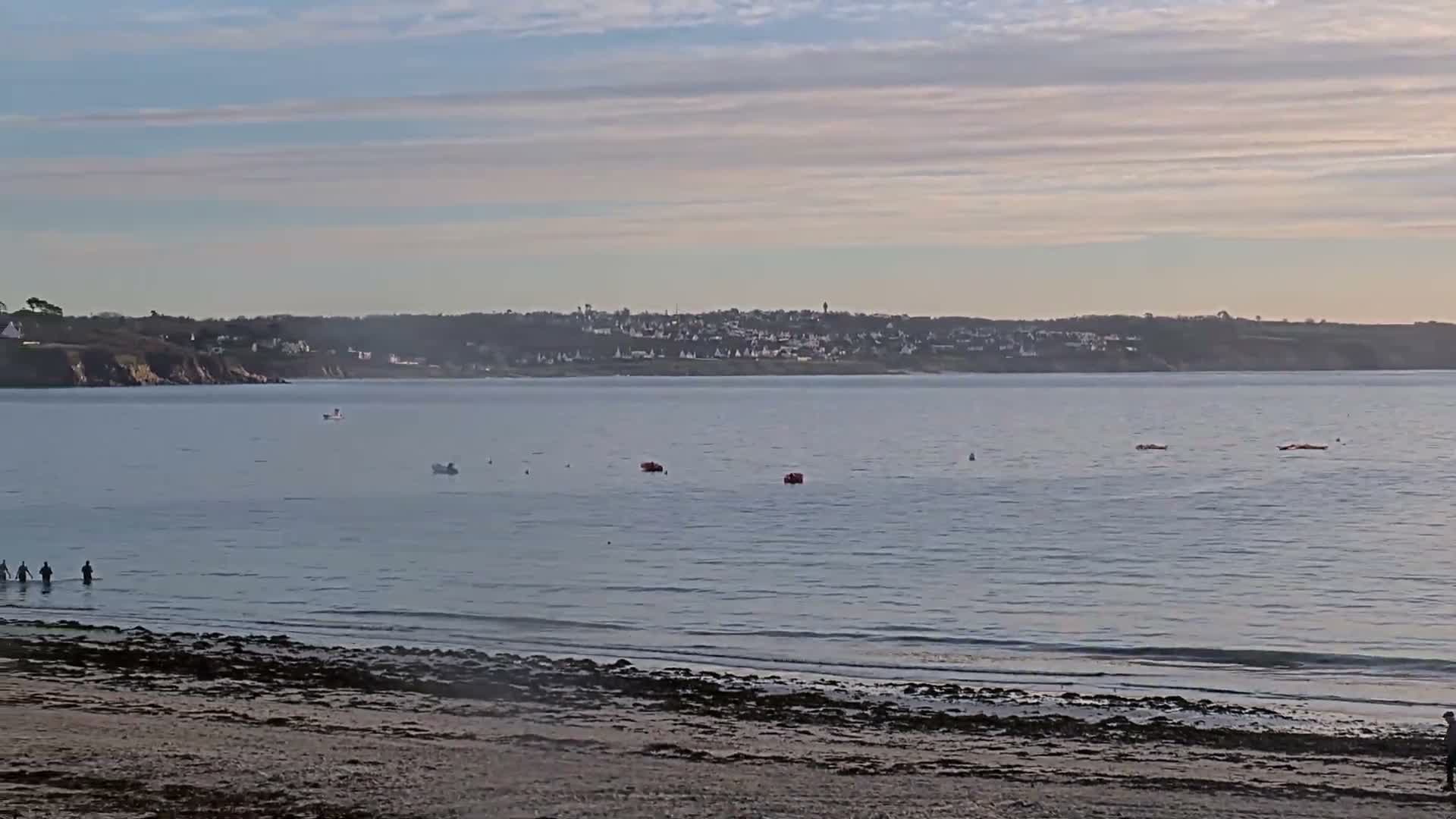 A calm bay with a sandy, seaweed-strewn beach in the foreground, where several people wade in the water, features distant boats and a coastal town under a partly cloudy sky.