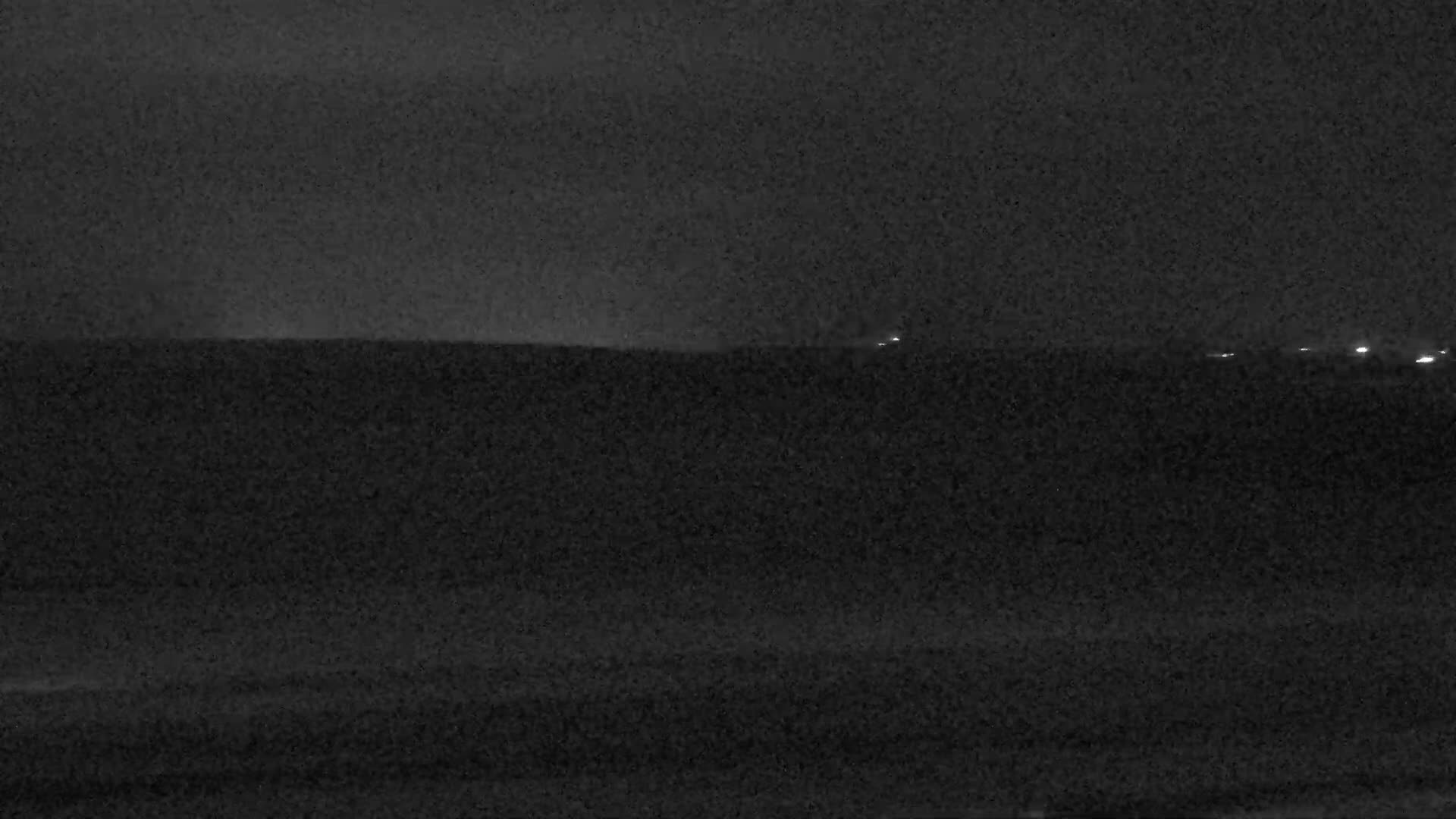 A very dark and grainy outdoor night scene shows a featureless foreground separated by a horizon from a distant area with several small, bright lights, under a dark sky.