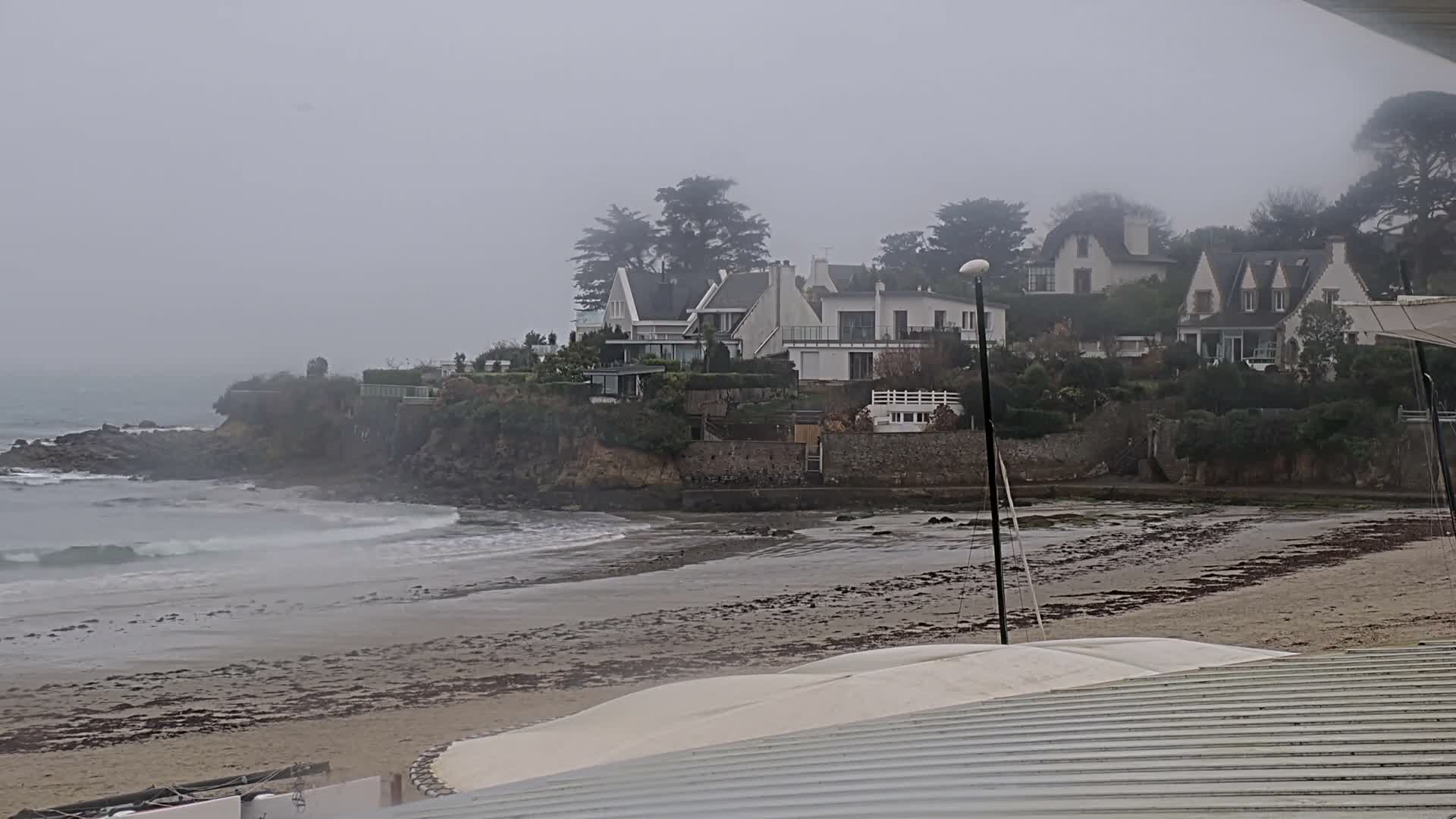 The image depicts an overcast and hazy coastal scene with waves breaking on a sandy beach scattered with seaweed, leading up to a rocky shore and several houses nestled among trees on a misty, distant hillside.