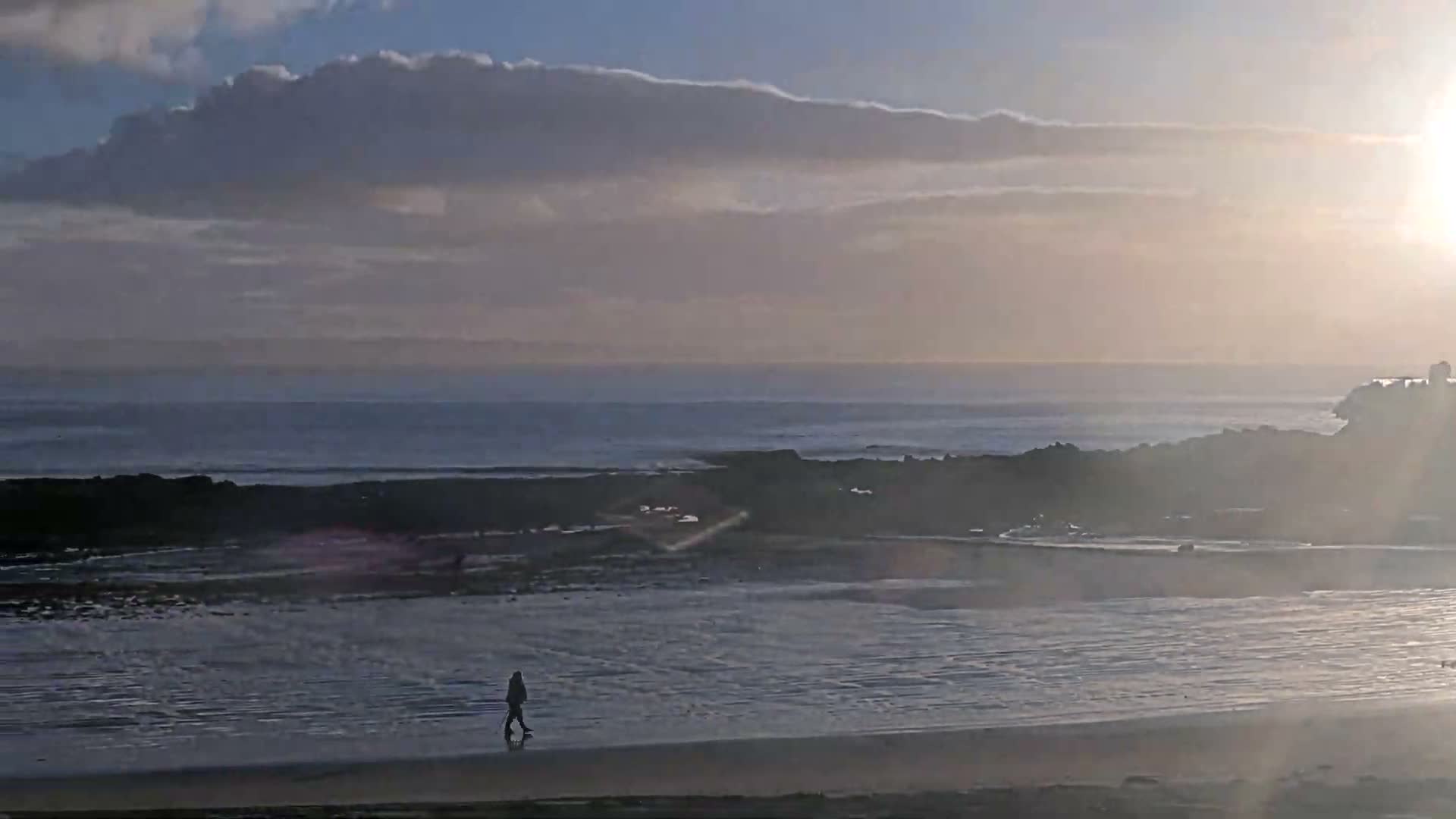 A lone person walks across a wet, reflective beach with a rocky coastline and ocean extending under a partly cloudy sky, with bright sunlight shining from the right.