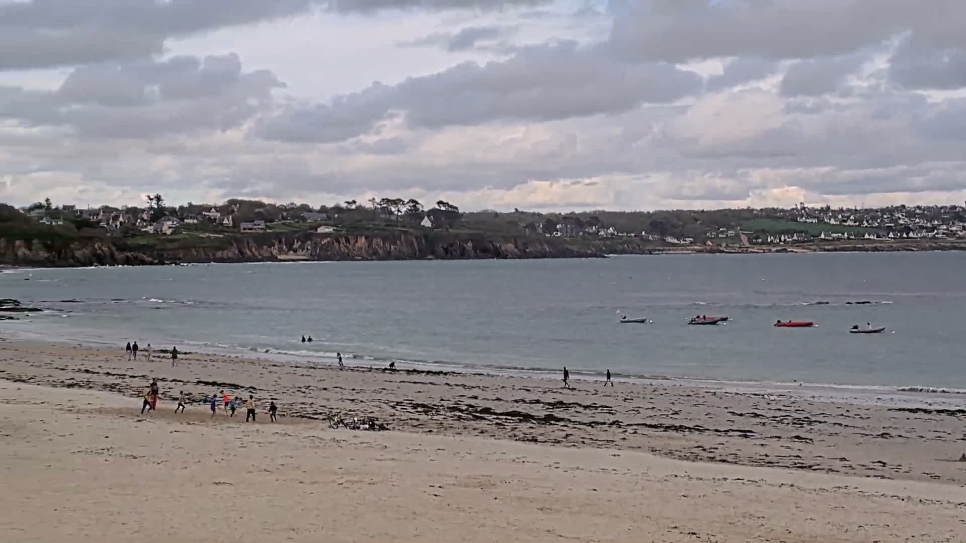 A lone person walks across a wet, reflective beach with a rocky coastline and ocean extending under a partly cloudy sky, with bright sunlight shining from the right.