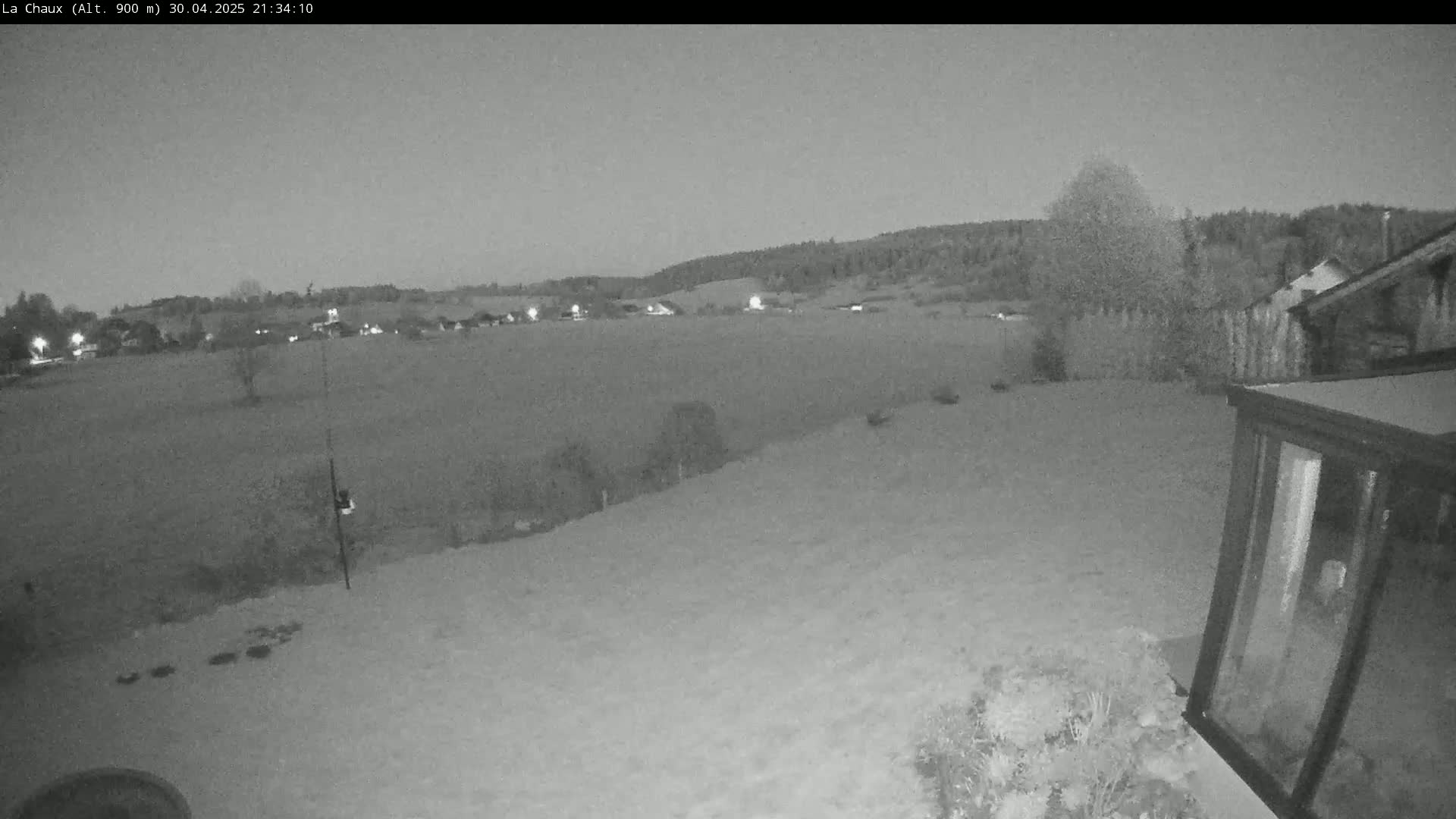 A nighttime, low-light view shows a flat, snow-covered field extending to a distant line of trees and houses under a dusky sky.