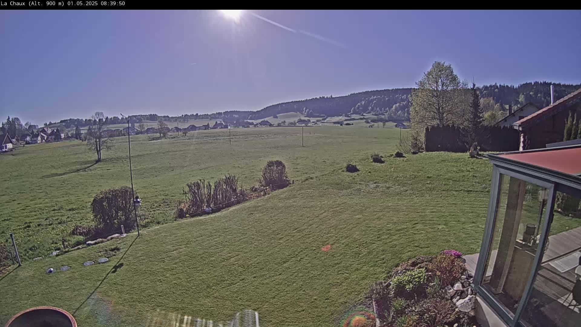 A sunny day reveals a sprawling green field, bordered by a small village and distant hills, partially obscured by a modern glass sunroom and a neatly kept garden.