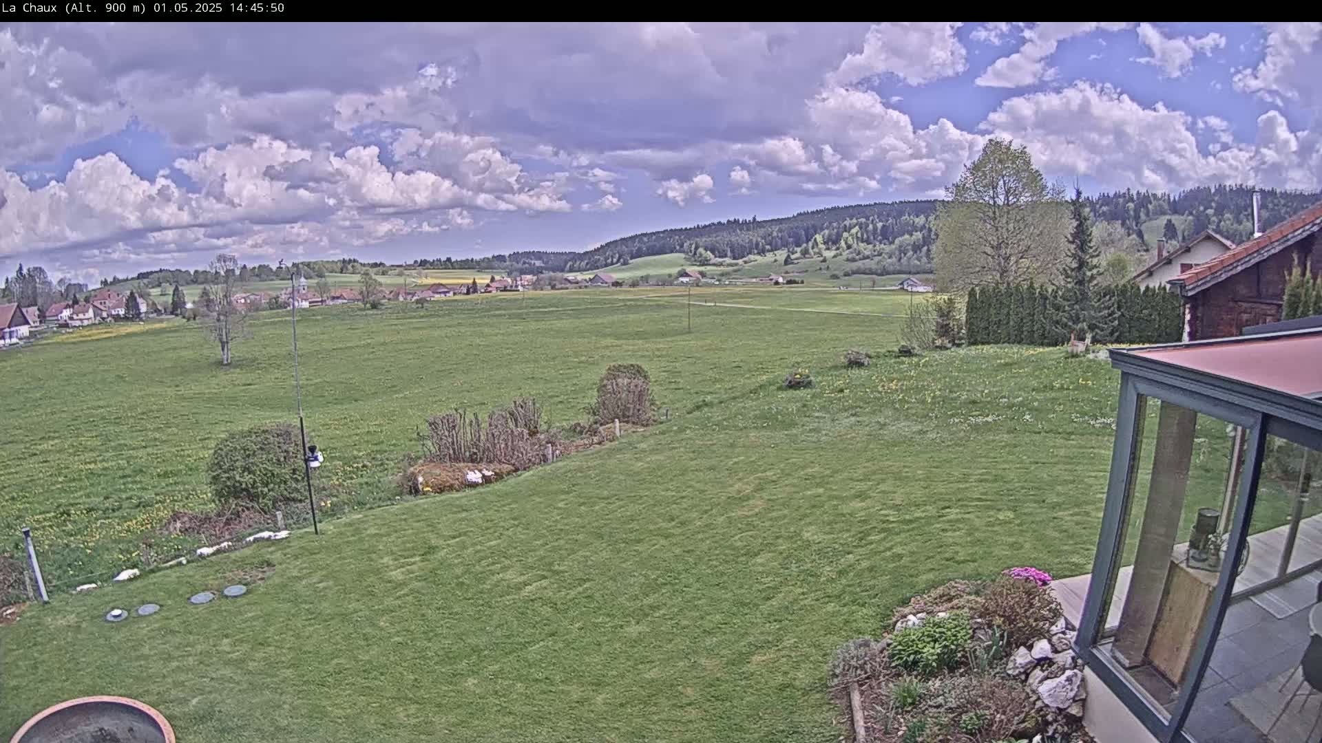 A partly cloudy sky overlooks a sprawling green lawn, a distant village, and a wooded hillside.