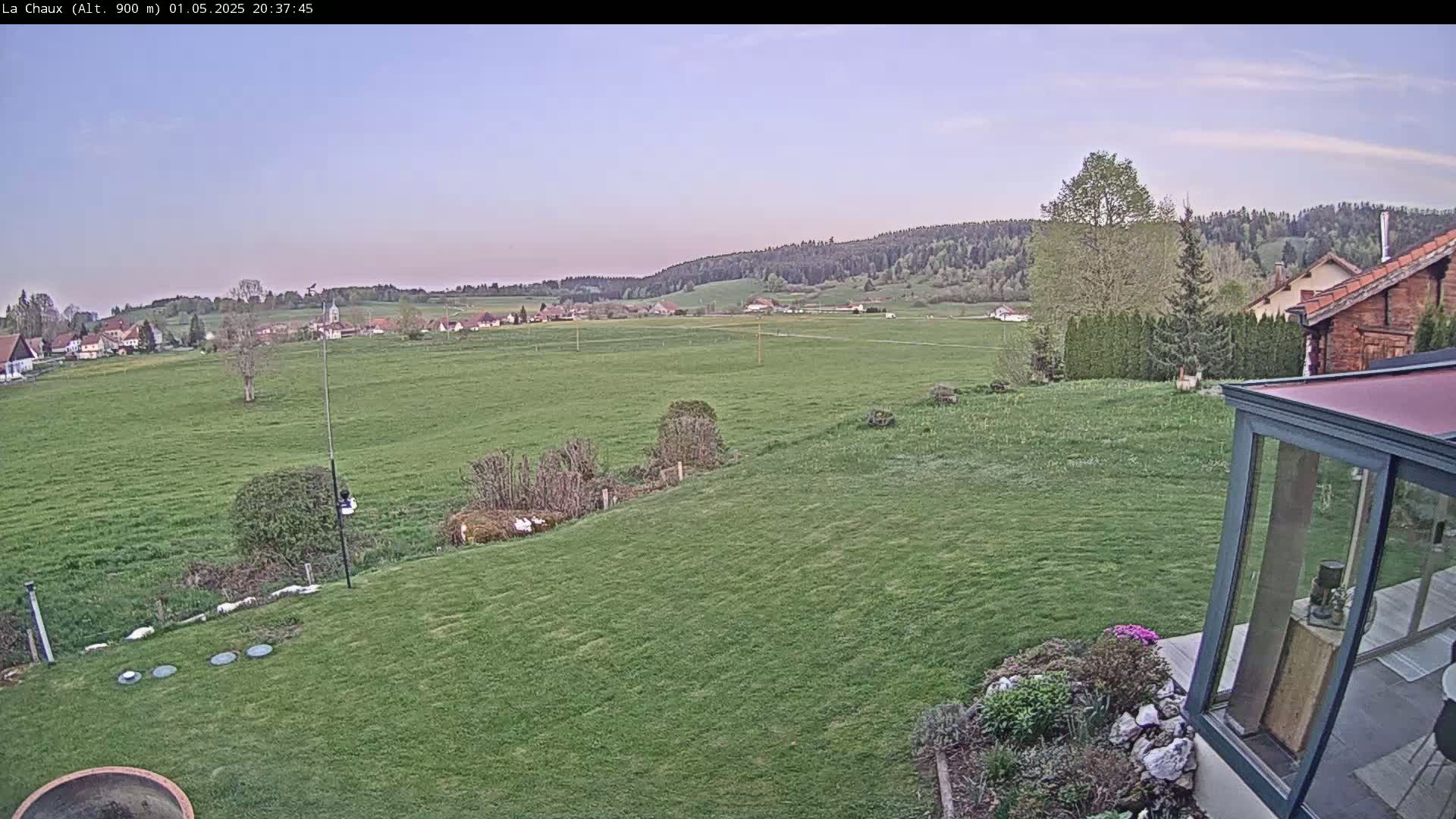 A grassy yard slopes down to a flat expanse of farmland, beyond which lies a village nestled against a hillside under a pale evening sky.