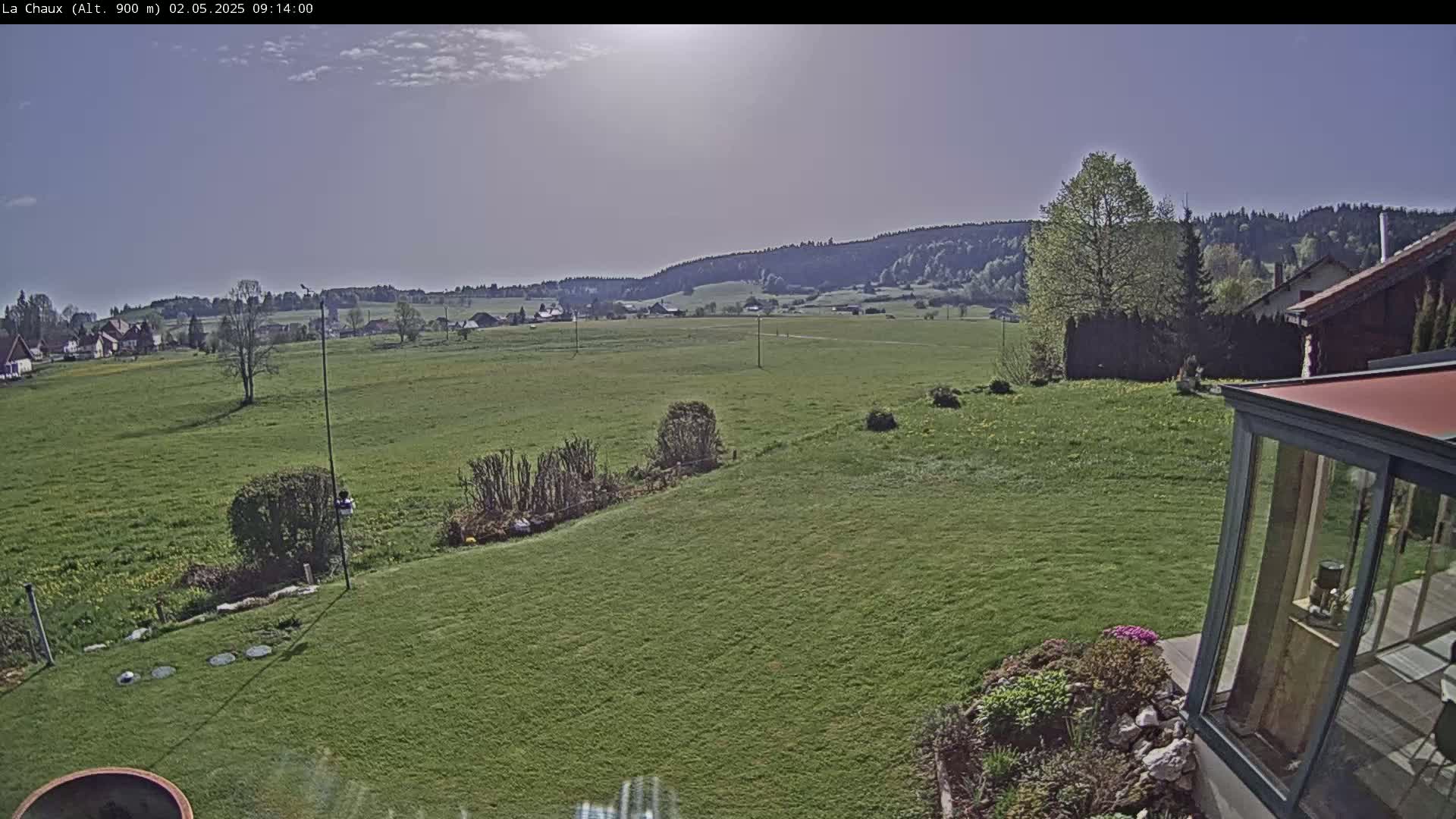 A sunny day reveals a sprawling green field, bordered by a few houses and trees, with a distant forested hillside under a mostly clear sky.