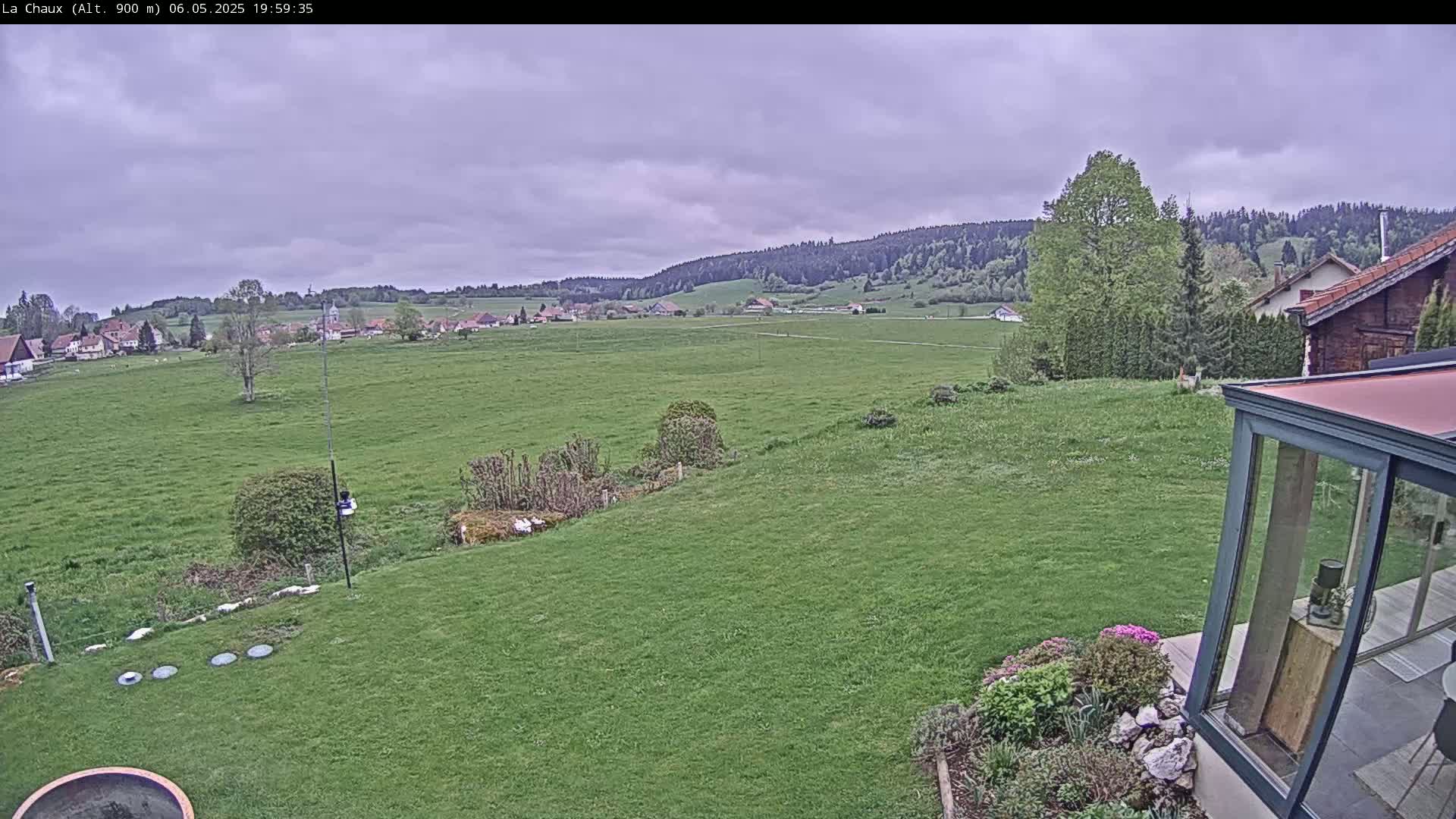 A verdant lawn slopes down to a vast green field, under a cloudy sky, with a village and distant hills visible in the background.