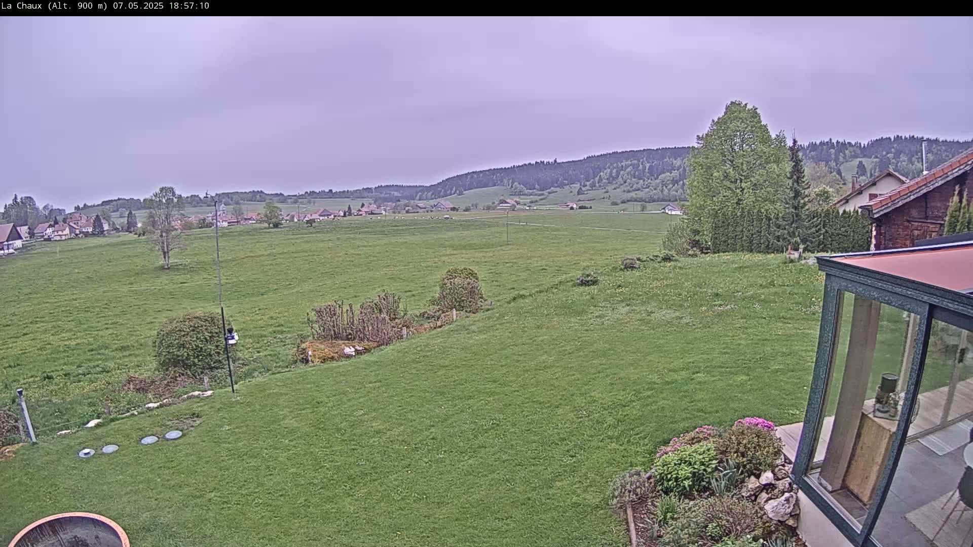 A grassy backyard slopes down to a field, beyond which a village and distant forested hills are visible under an overcast sky.