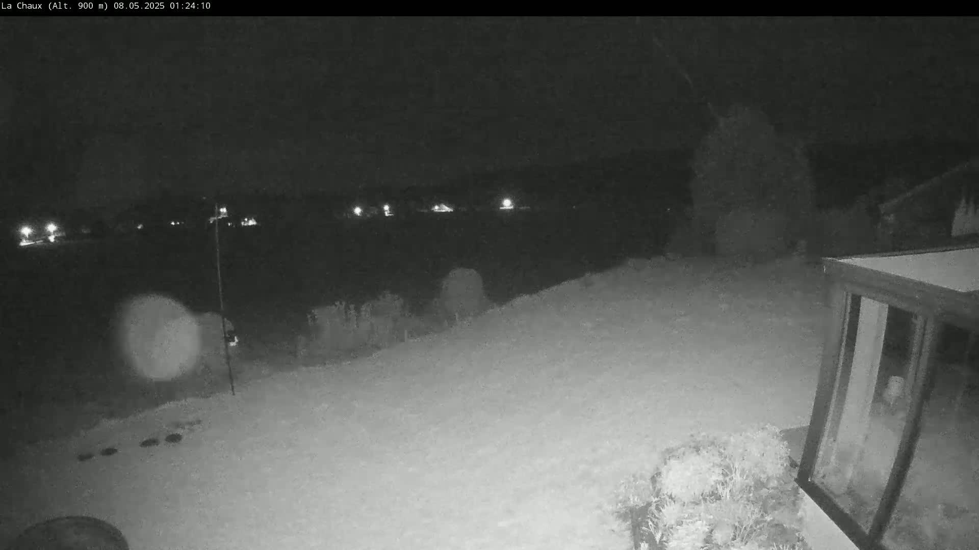 A snow-covered backyard at night is illuminated by distant lights, showing a portion of a structure with a glass door in the lower right corner.