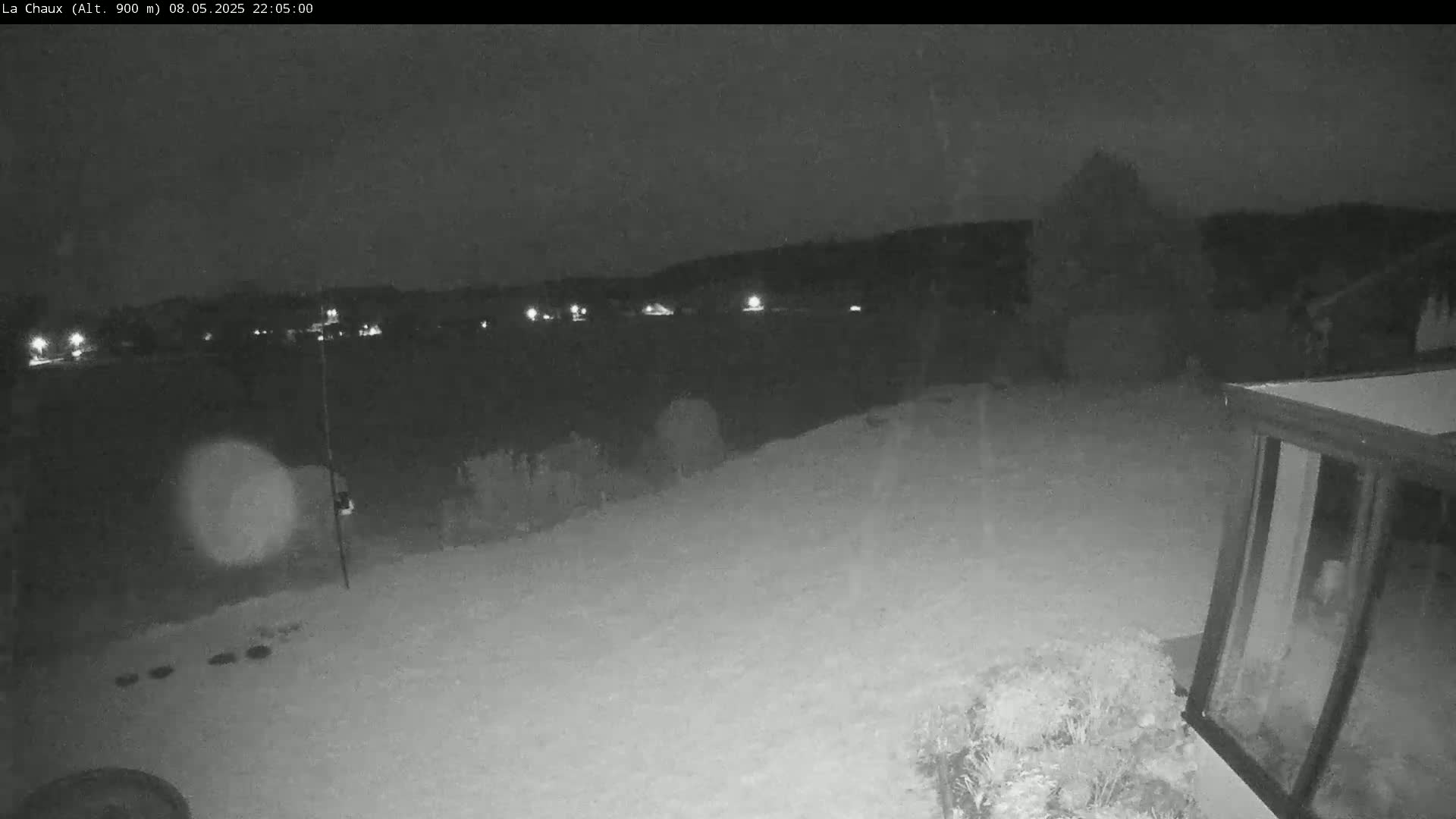 A nighttime, snowy backyard scene shows a distant line of lights, a dark building, and some shrubs, under a dark sky.