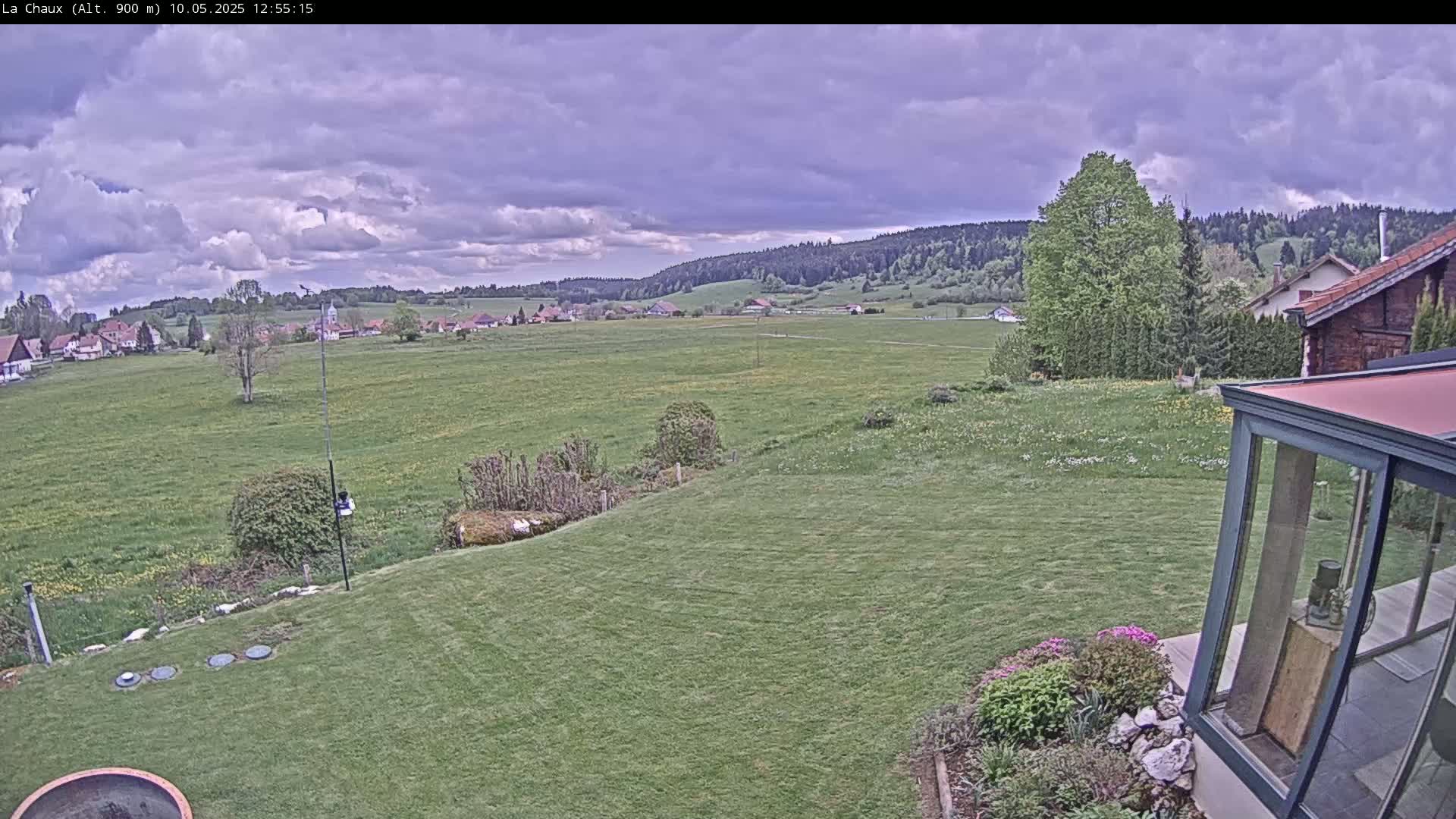 A sprawling green lawn slopes gently downward toward a village nestled in a valley under a partly cloudy sky.