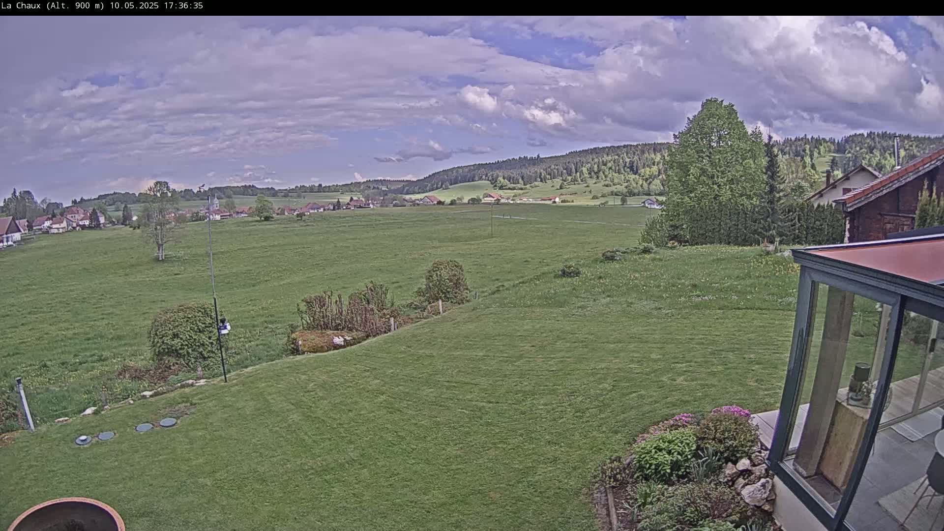 A partly cloudy sky overlooks a sprawling green field, a village in the distance, and a grassy yard leading to a modern glass sunroom.