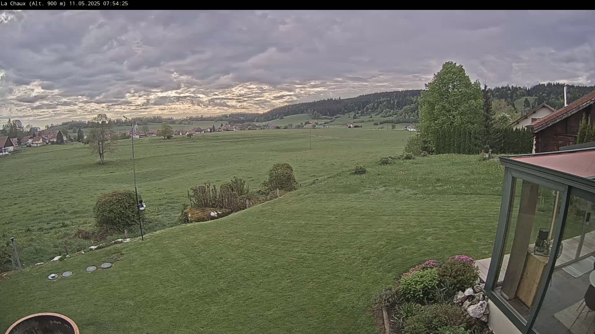 A verdant lawn slopes down to a broad, grassy field under a mostly cloudy sky, with a village and distant hills visible in the background.