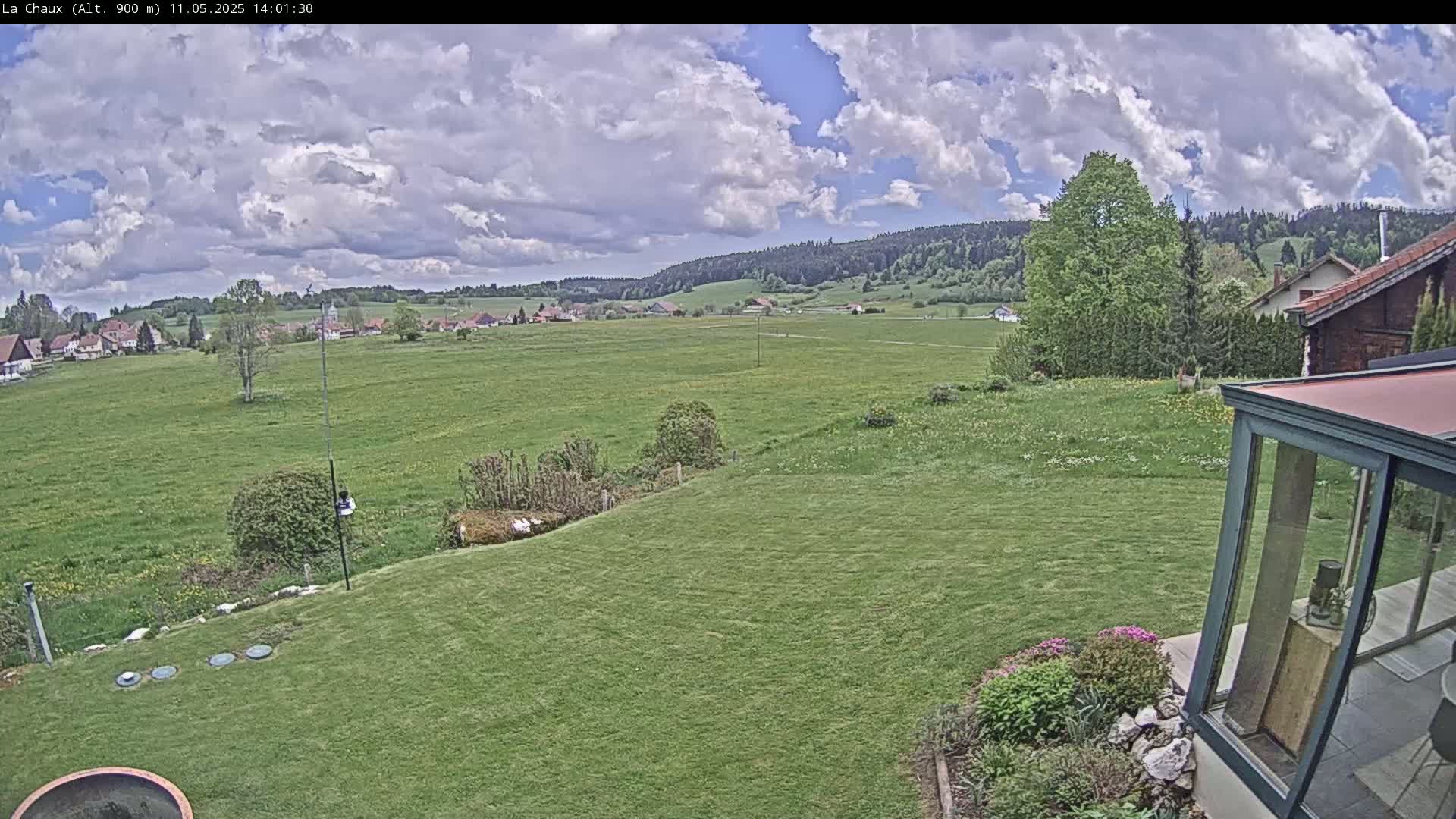 A partly cloudy sky overlooks a verdant landscape encompassing a grassy yard, a distant village, and a forested hillside.