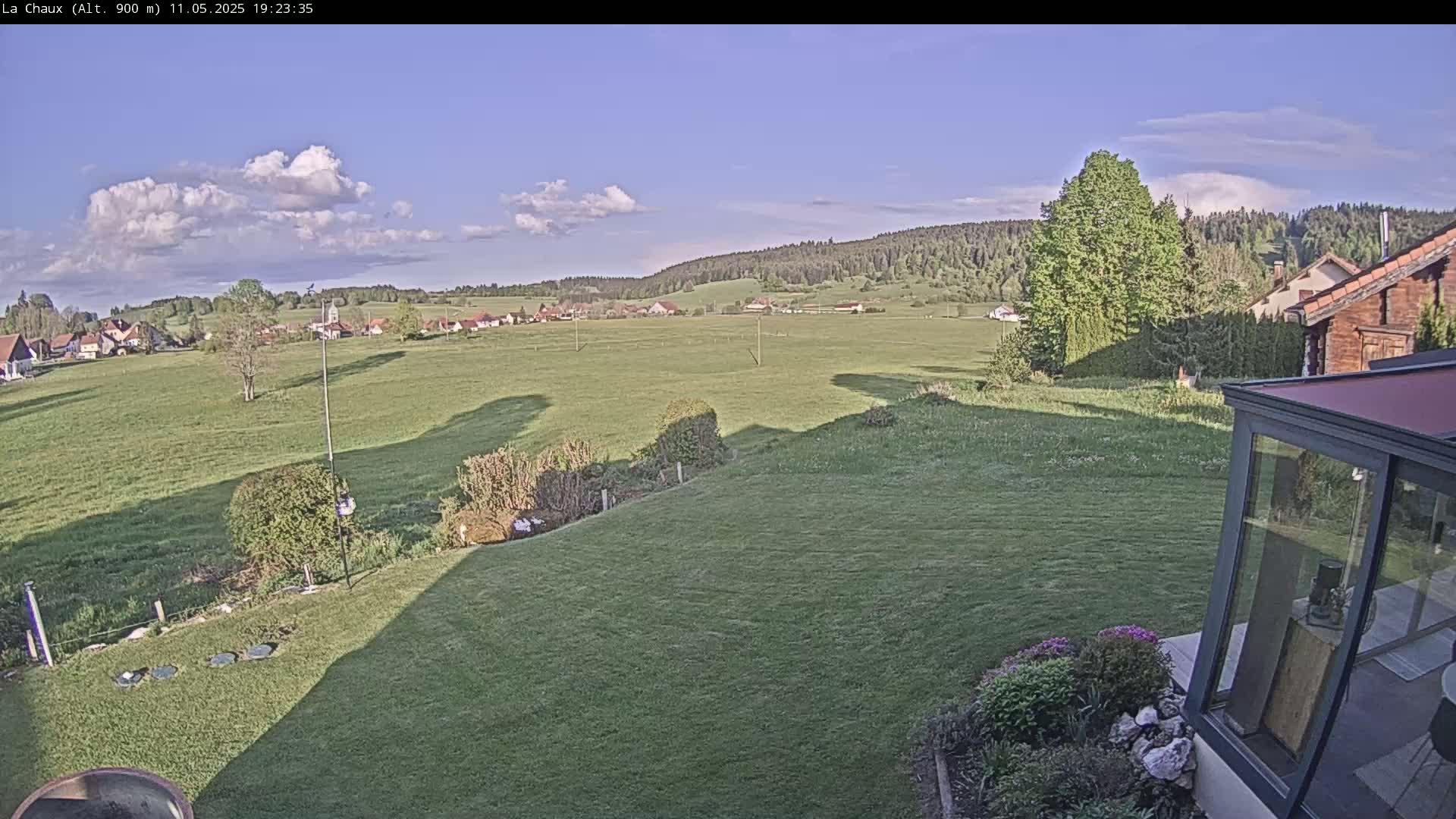 A mostly sunny day shows a green grassy yard sloping down to a flat field with a small village and forest in the distance.