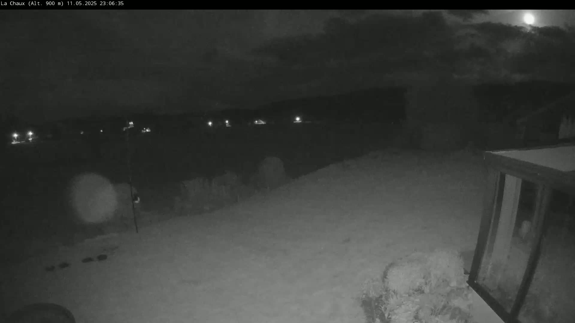 A nighttime, low-light outdoor scene shows a partially cloudy sky with a bright moon, a snow-covered ground, and a dark structure in the lower right corner.