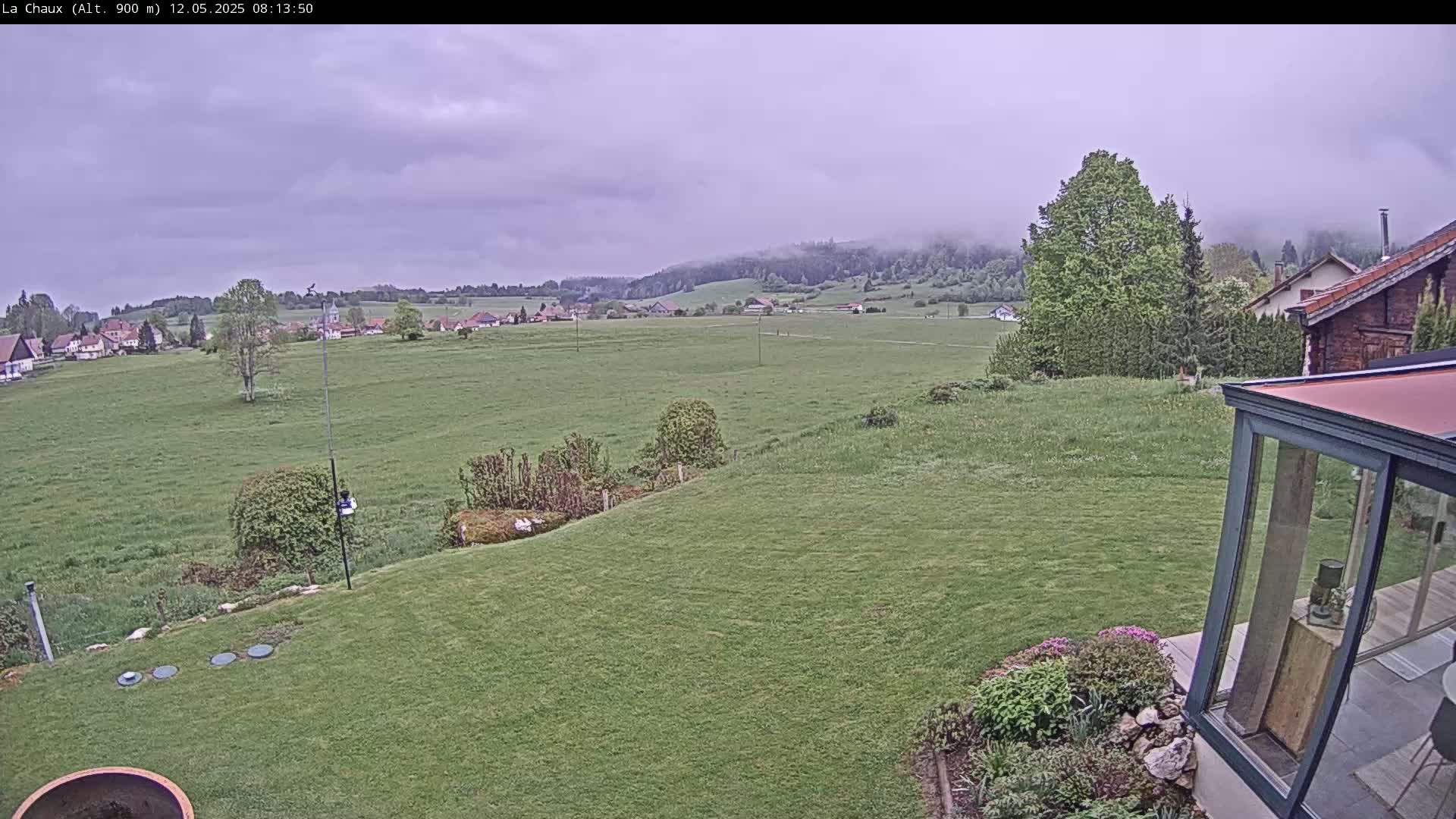 A sprawling green lawn slopes down to a distant village nestled in a valley under a cloudy sky.