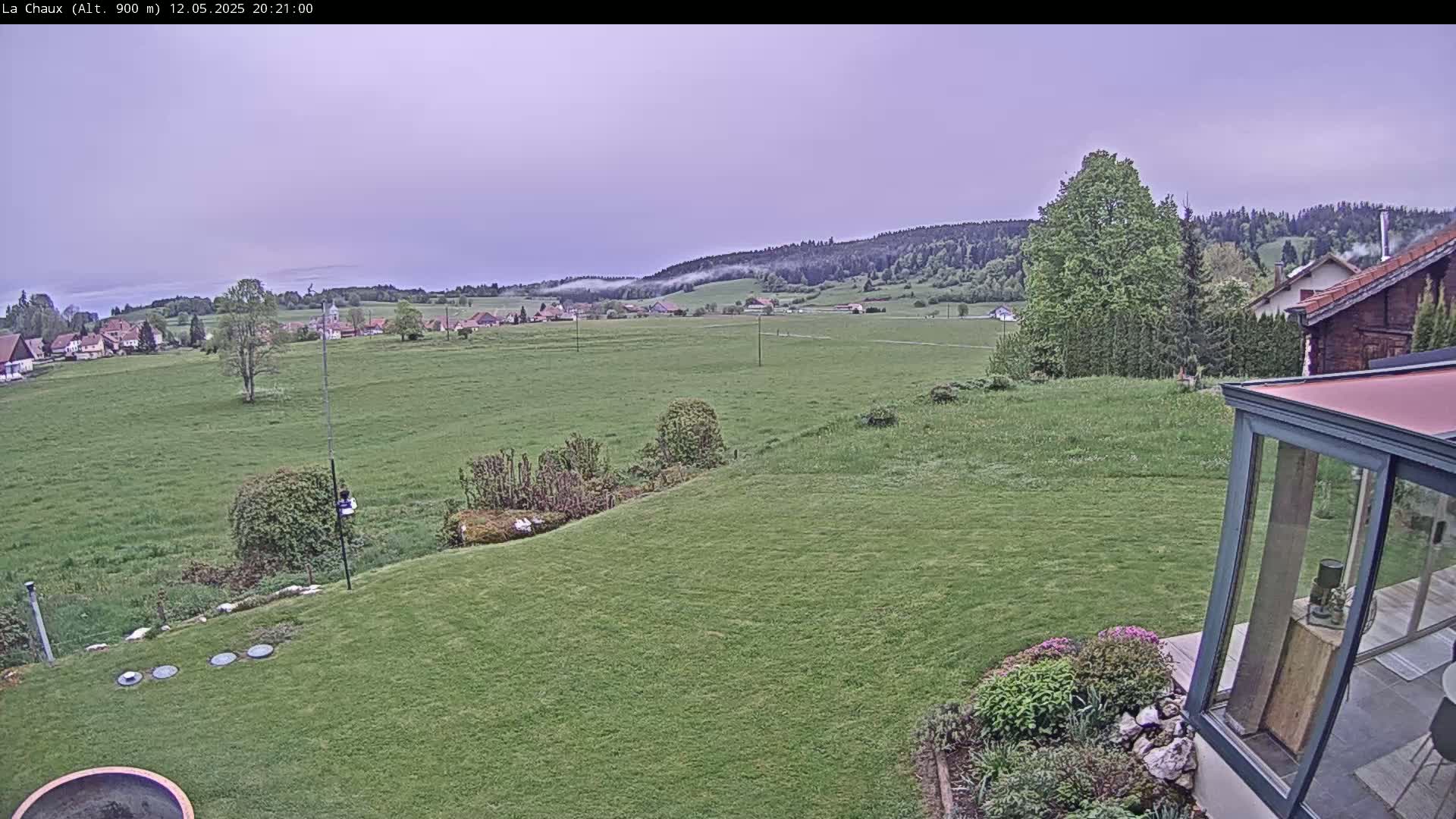A grassy backyard slopes down to a field, beyond which lies a village nestled against a hazy, forested hillside under an overcast sky.