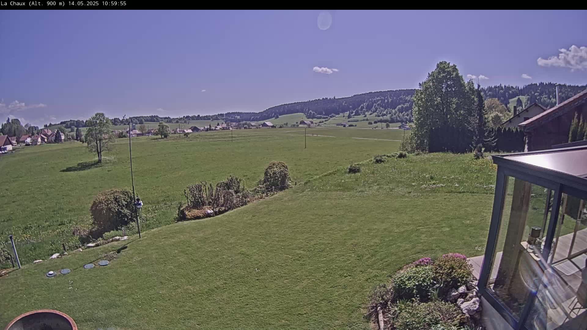 A sunny day reveals a sprawling green lawn leading to a village nestled in a valley, backed by a line of dark green hills under a mostly clear blue sky.