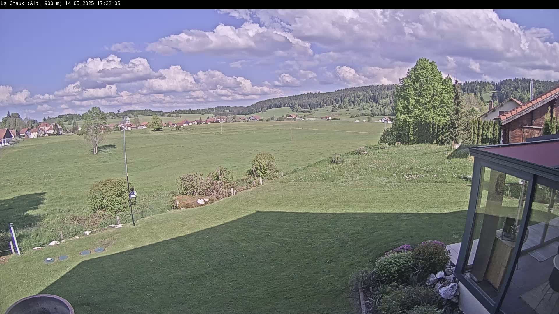 A partly sunny day reveals a sprawling green field with a village in the distance, nestled against a backdrop of dark green hills, all viewed from a garden with a sunroom in the foreground.