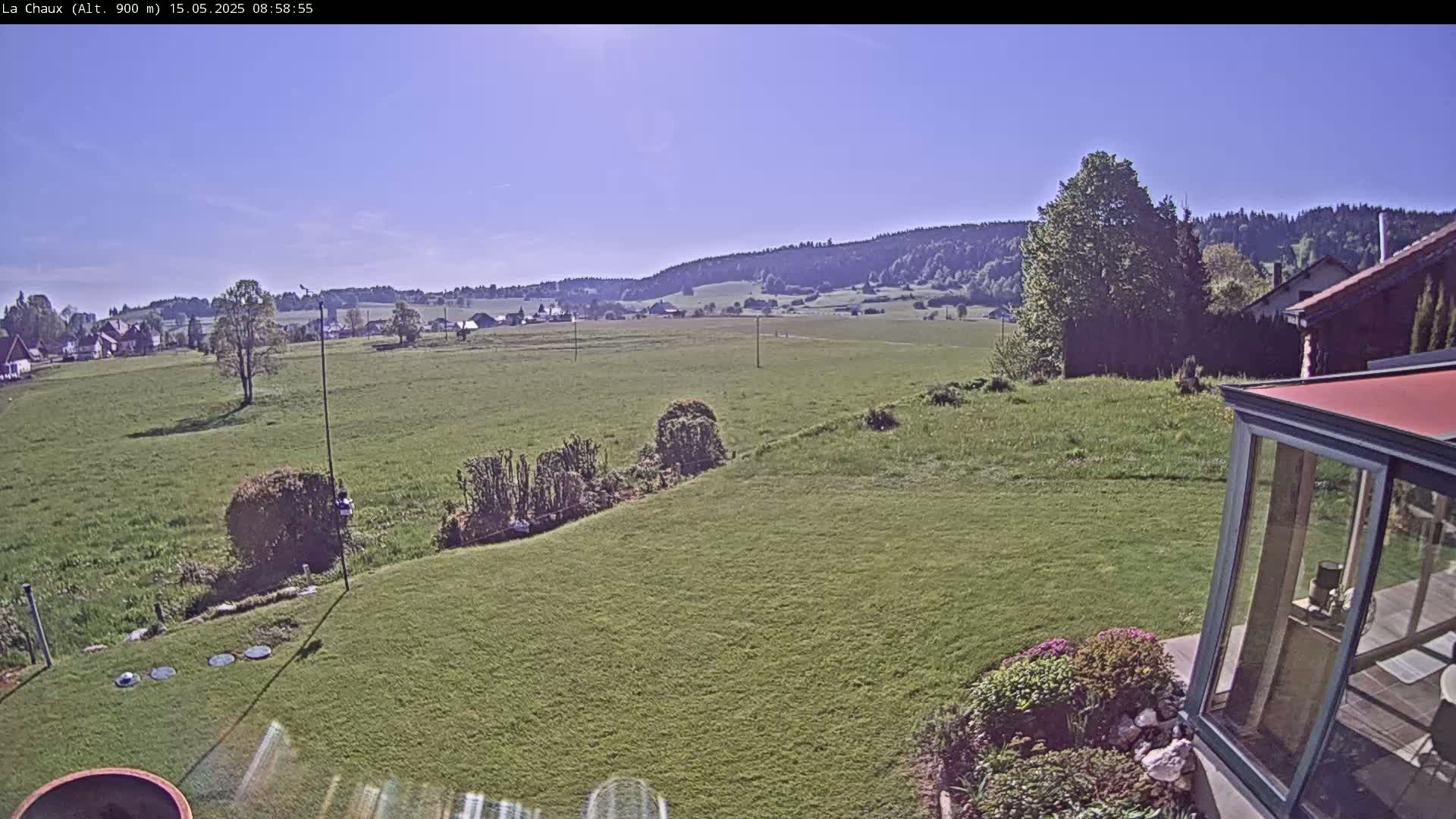 A grassy backyard scene on a sunny day, overlooking a verdant field and distant hills with a few houses and trees visible in the background.