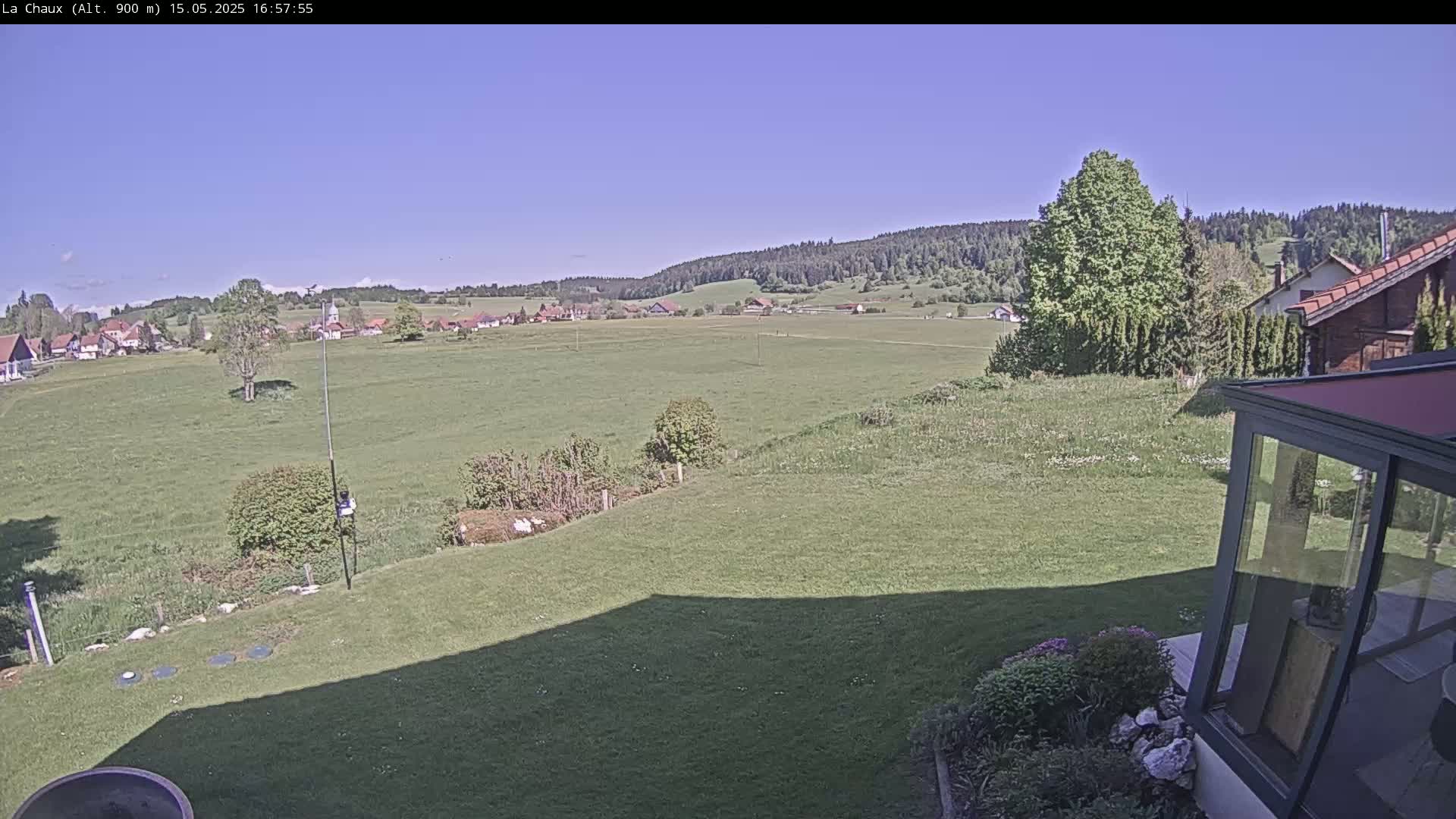 A sprawling green field stretches out to a village nestled against a forested hillside under a clear blue sky.