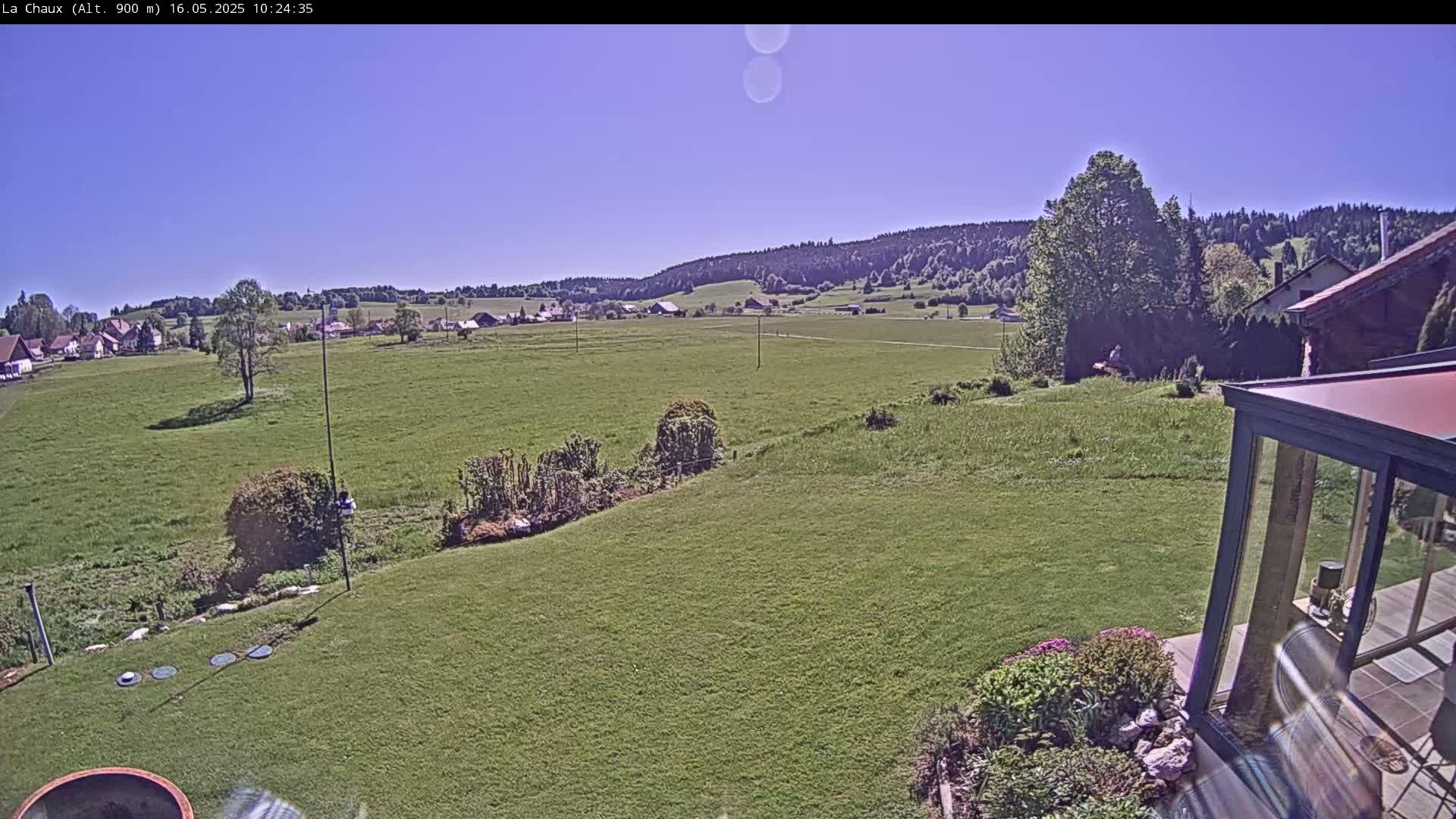 A sunny day reveals a sprawling green field, a small village in the distance, and a glass conservatory next to a lush garden.