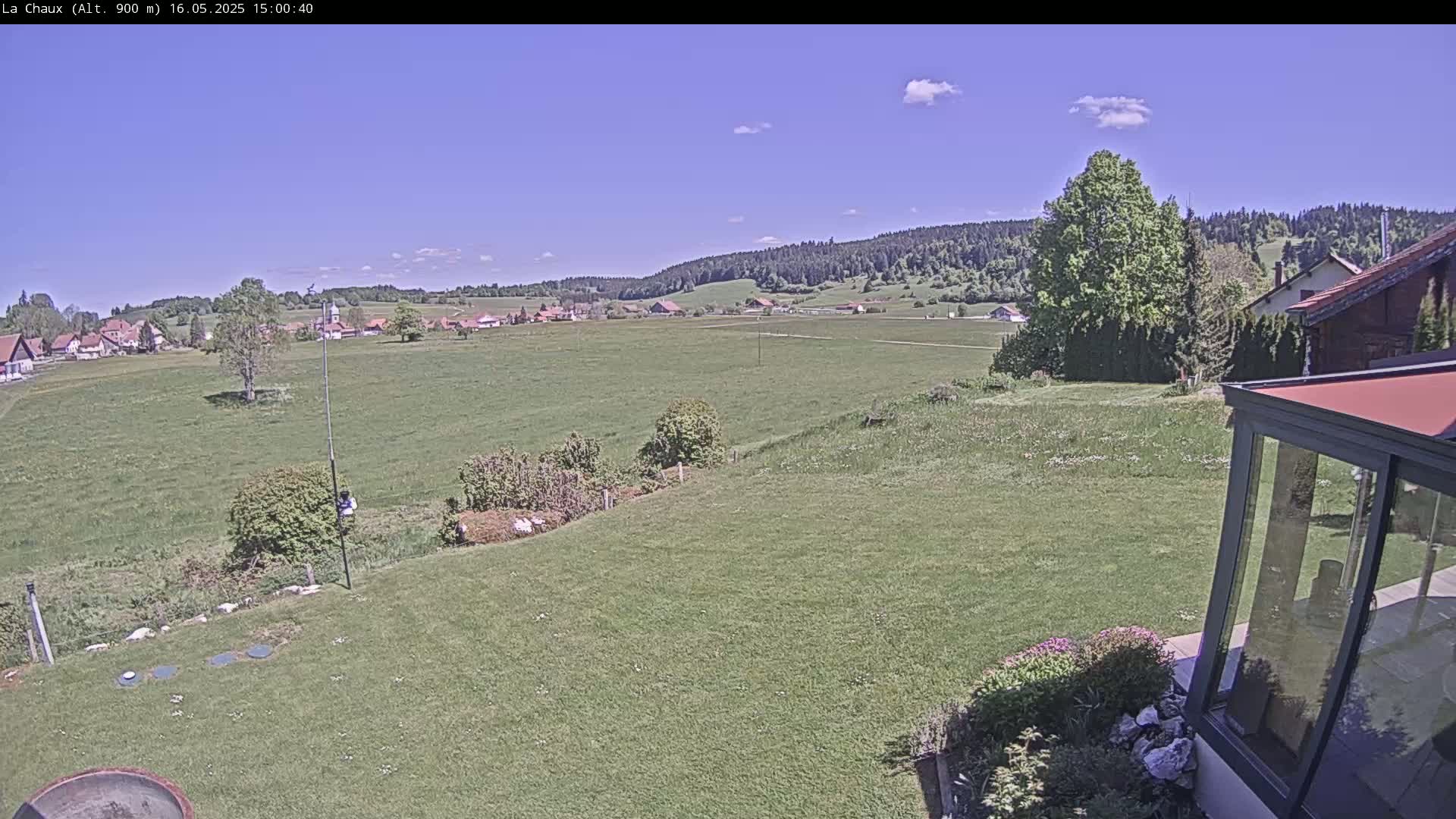 A mostly sunny day reveals a verdant field stretching towards a small village nestled at the foot of forested hills.