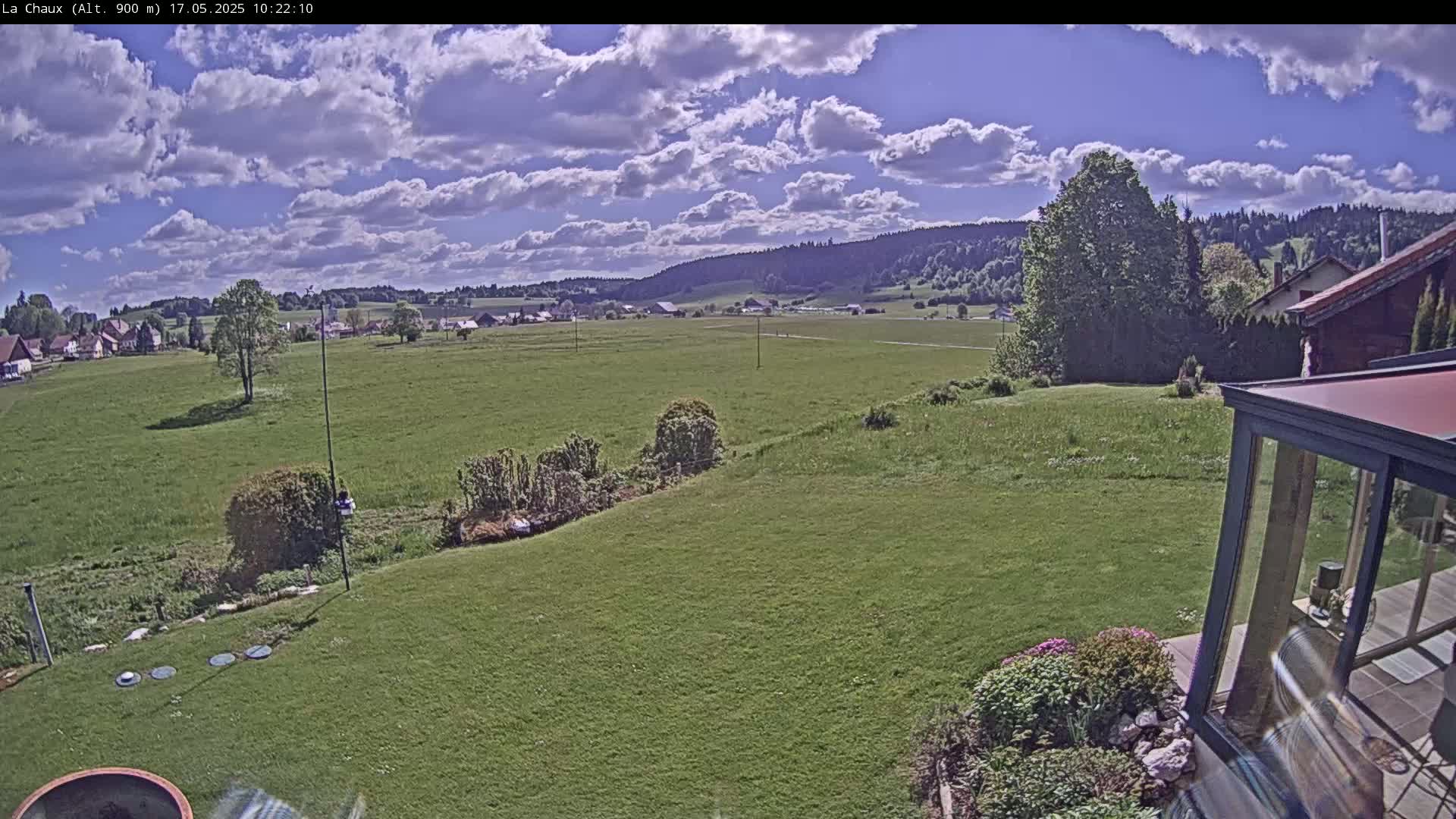 A partly cloudy sky overlooks a verdant field, a small village, and forested hills in the distance, with a portion of a backyard and modern glass structure visible in the foreground.